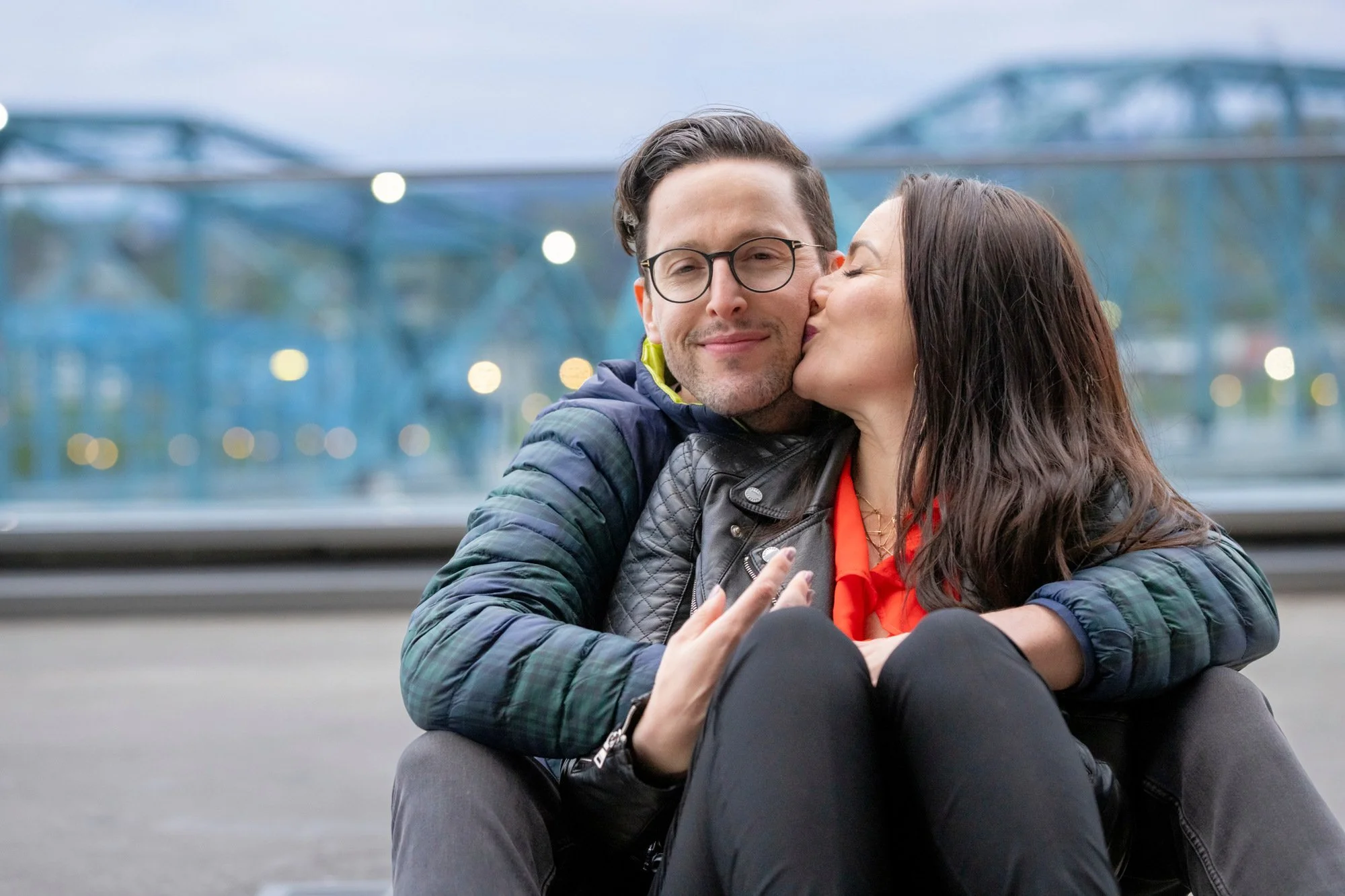 A man and woman sitting outdoors on a concrete surface with a blurred blue bridge and lights in the background. The woman is kissing the man on the cheek and they are both smiling. The man is wearing glasses and a blue jacket, and the woman has long 