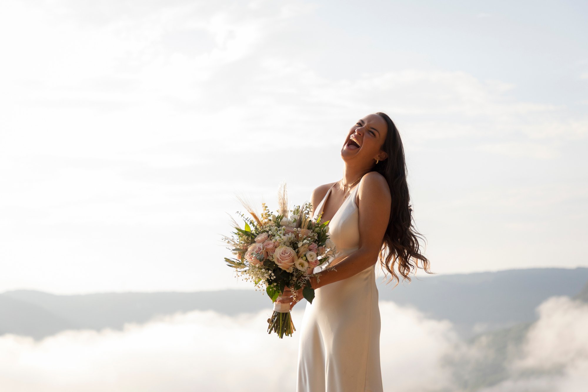 A woman in a white dress holding a bouquet of pink and white flowers, smiling and laughing outdoors against a cloudy sky and distant mountains.