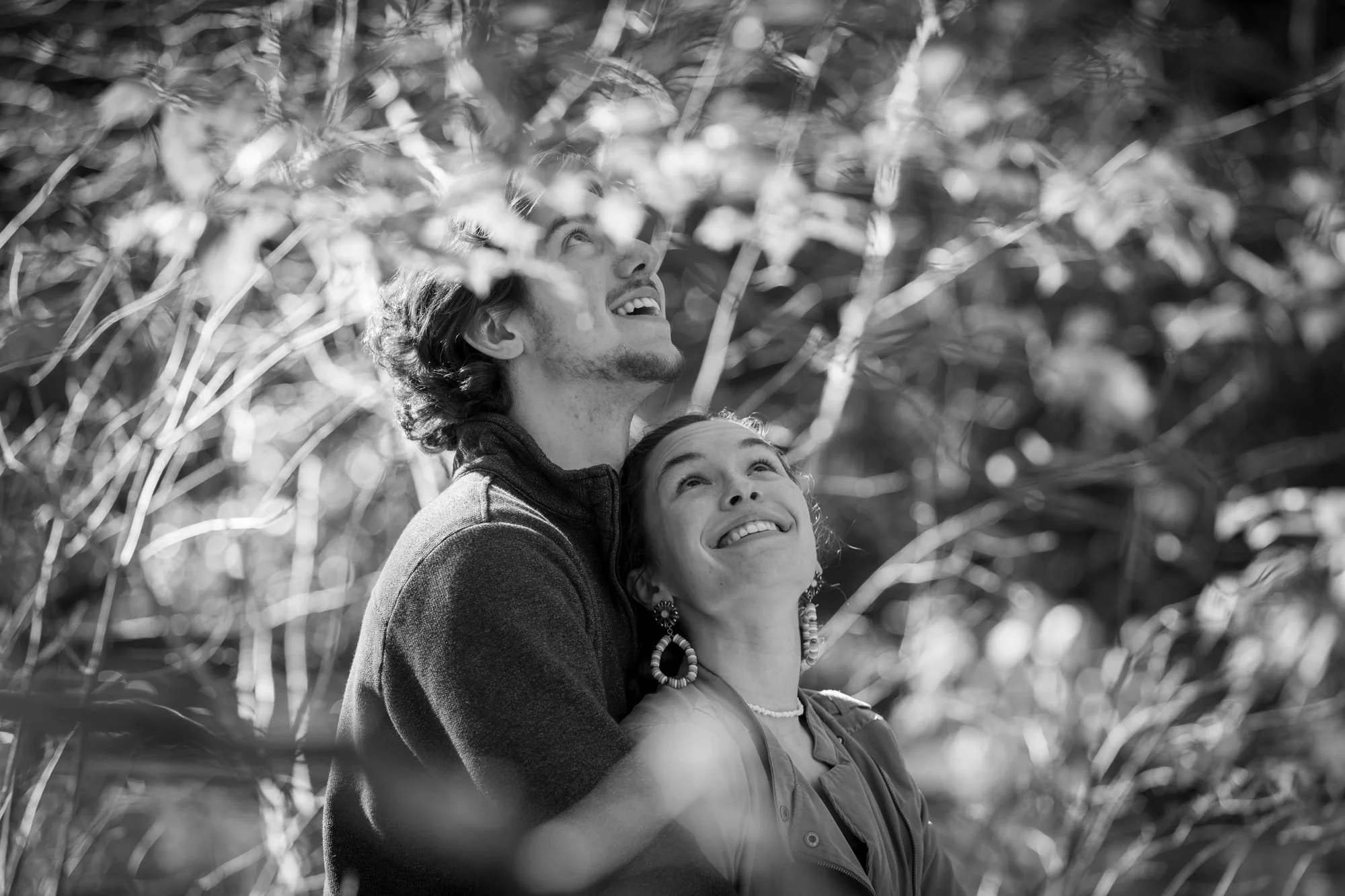 A man and woman cross-eyed looking up with smiles, surrounded by tall grasses or plants, in black and white.