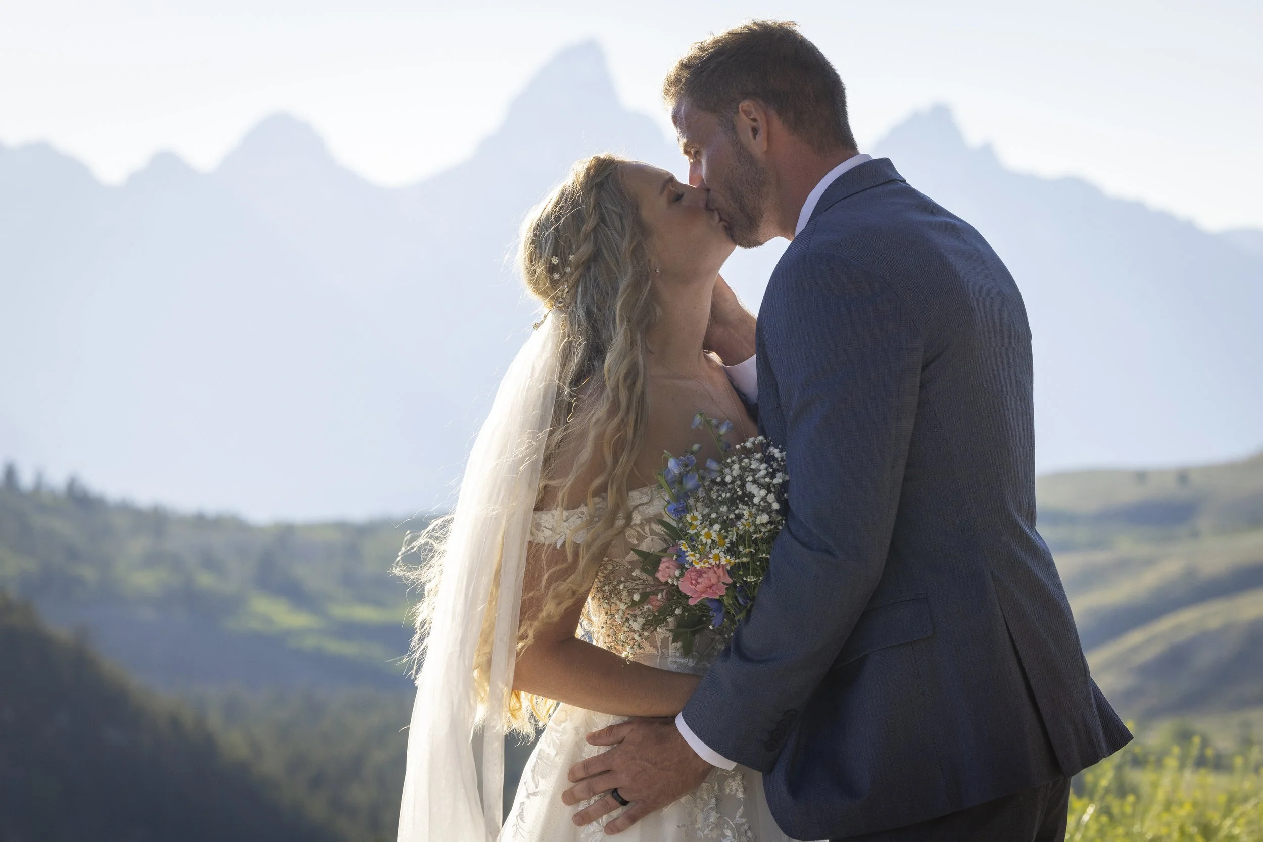A bride and groom sharing a kiss outdoors during their wedding, with mountains in the background.