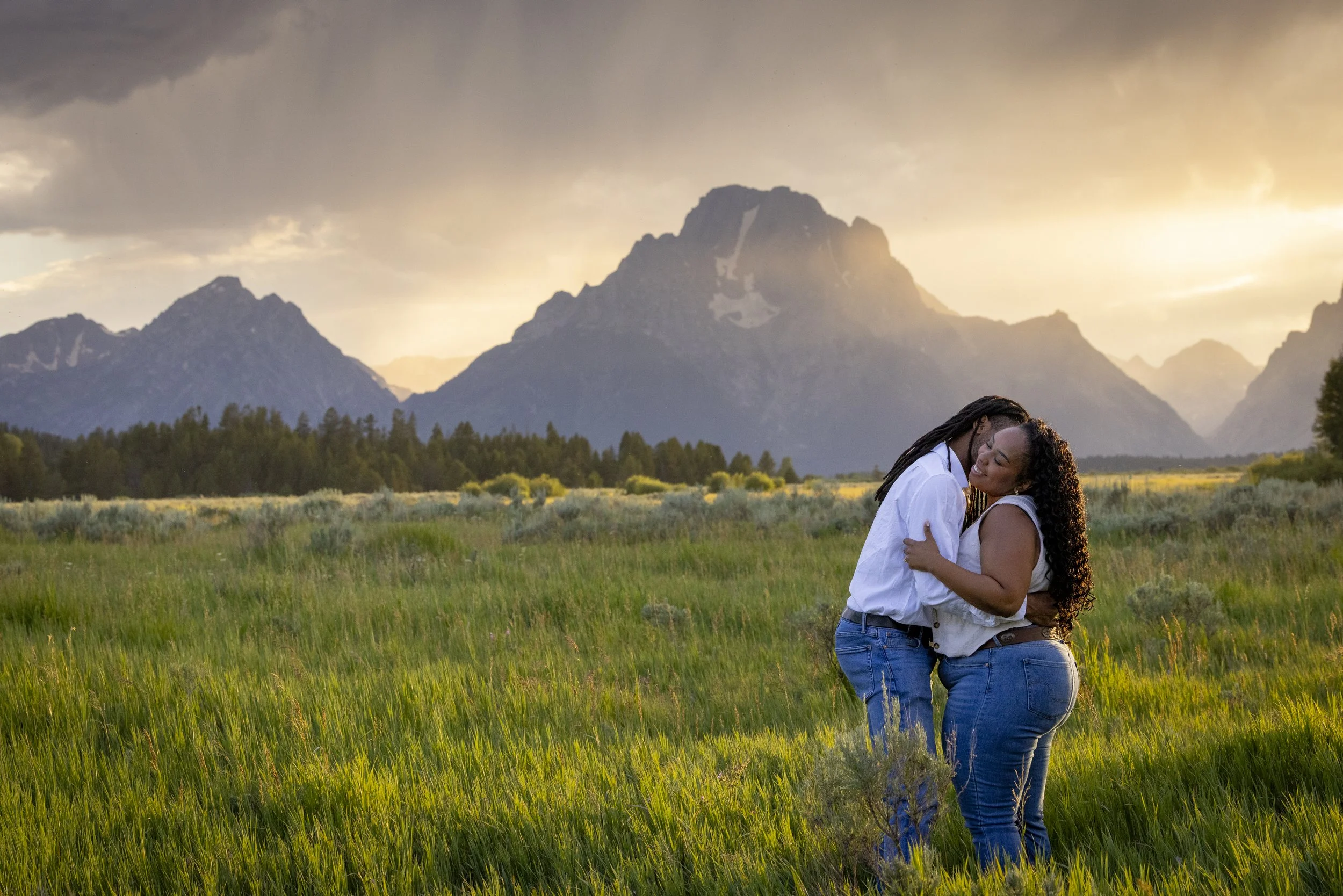 A couple embraces and kisses in a grassy field with mountains in the background during sunset.
