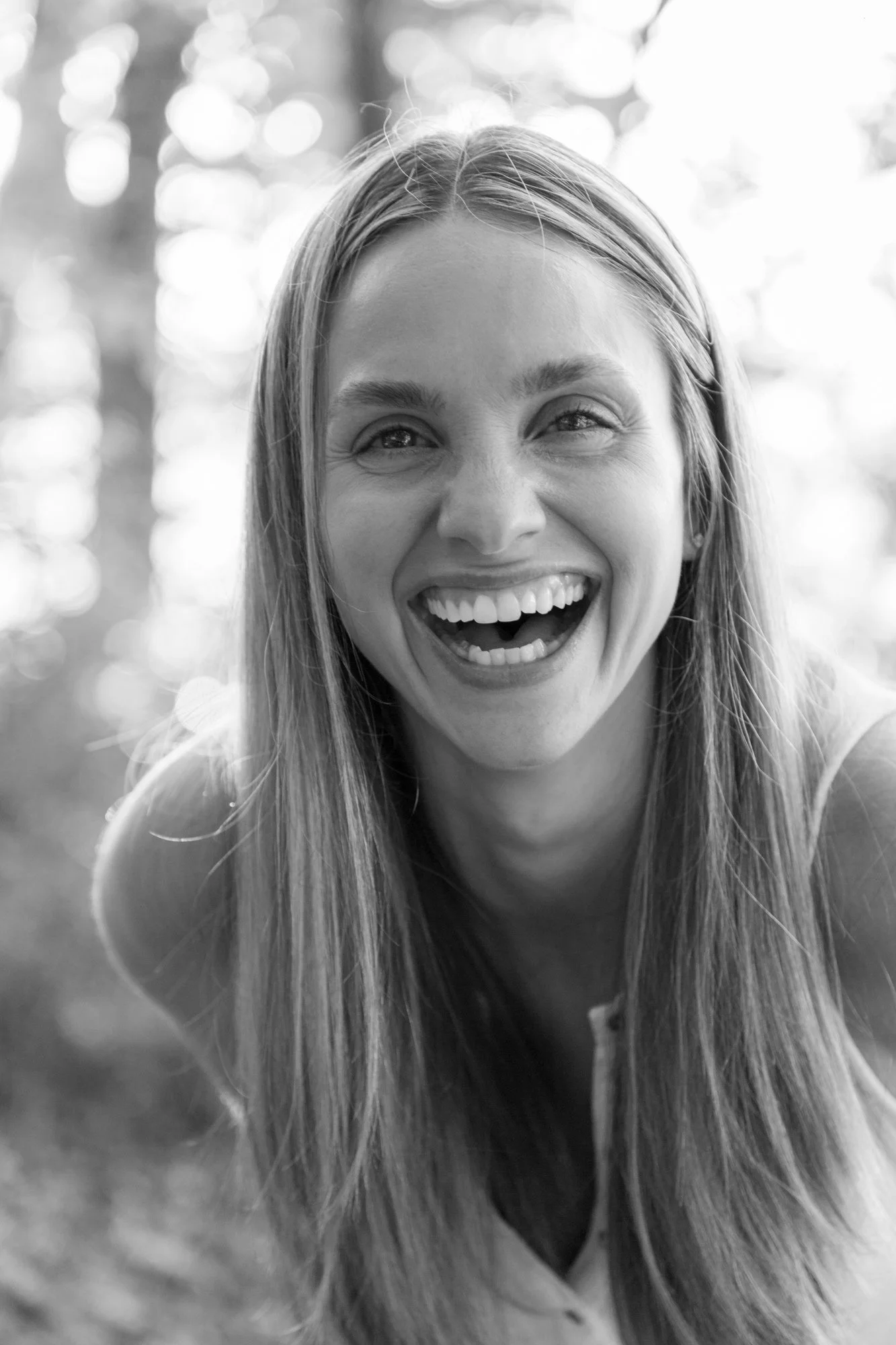 A black and white photo of a woman laughing outdoors, with trees and light in the background.
