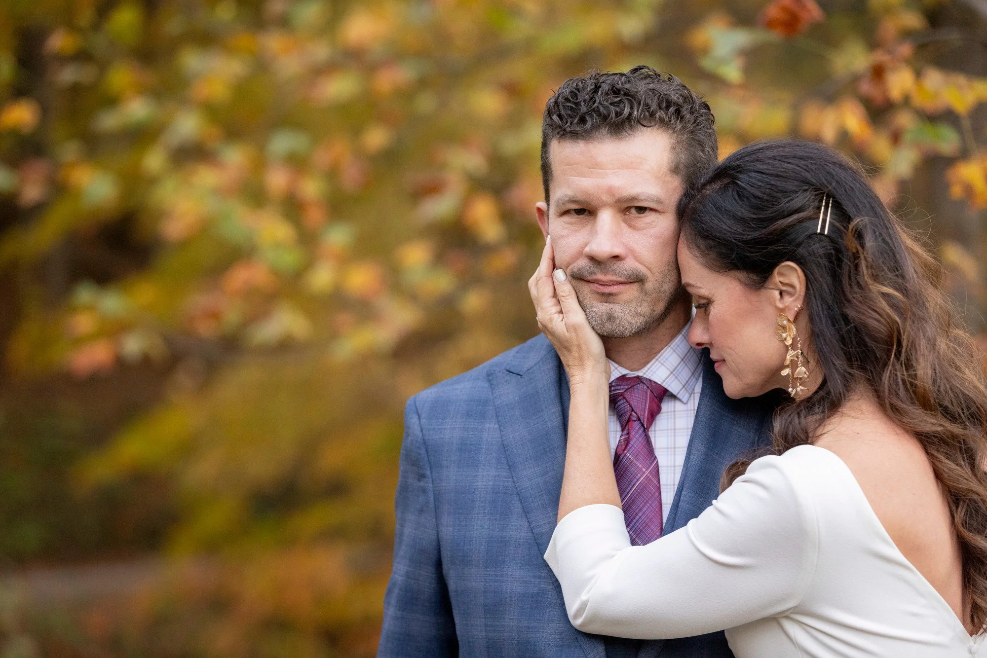 A man in a blue suit with a woman with dark hair and a white dress in an outdoor setting with autumn-colored trees, embracing affectionately.