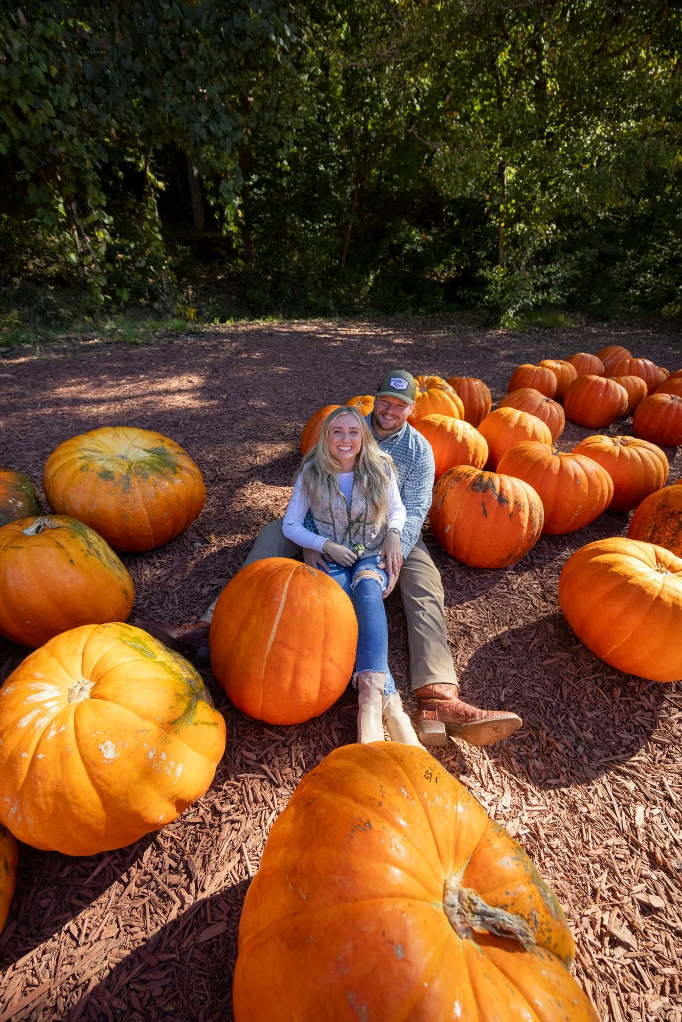 A smiling couple sitting on the ground surrounded by large orange pumpkins on a pumpkin patch with trees in the background.