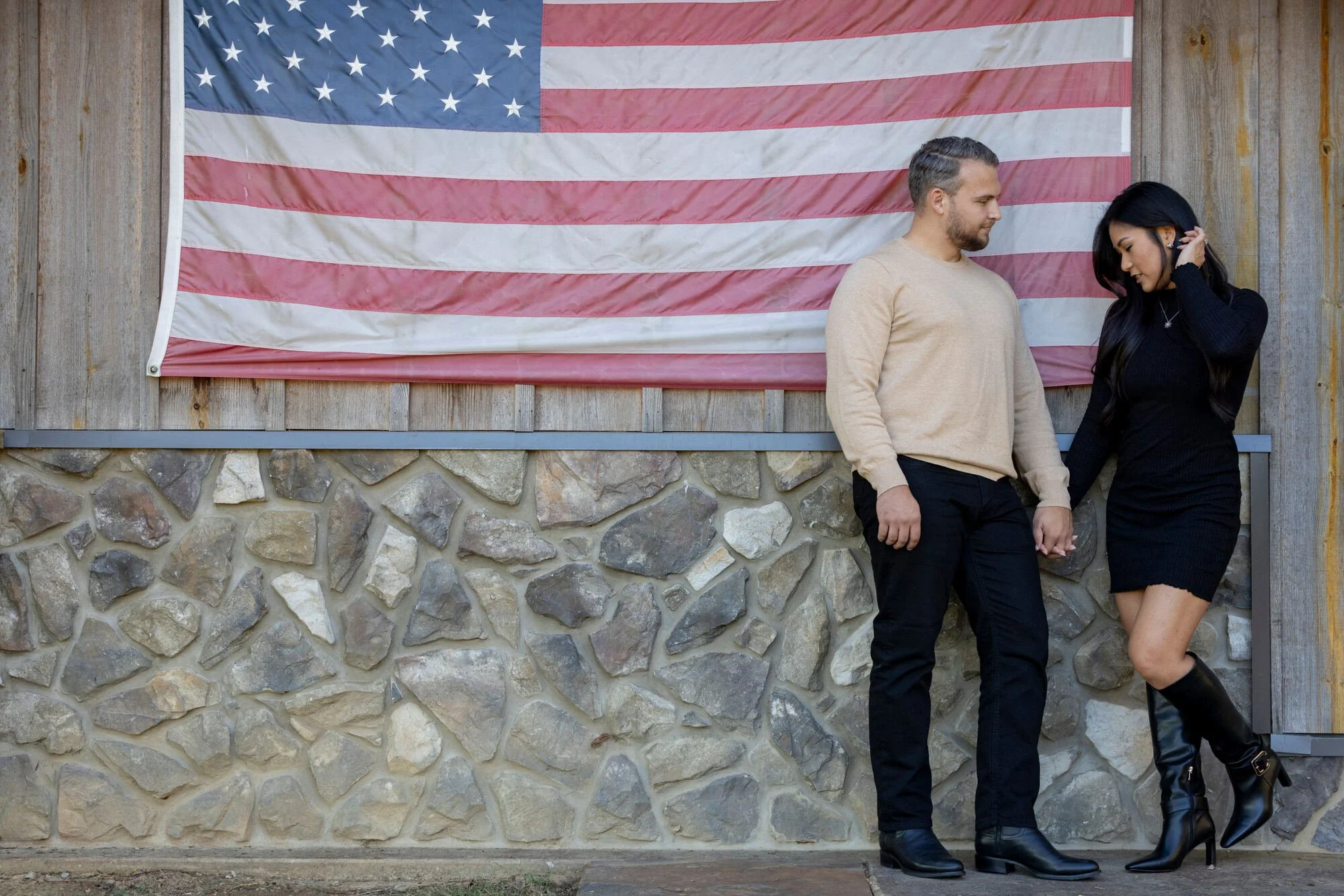 A man and woman standing close together outdoors in front of a large American flag on a wooden wall. The man has short hair, a beard, and is wearing a beige sweater and black pants. The woman has long dark hair, is dressed in a black outfit with a sh