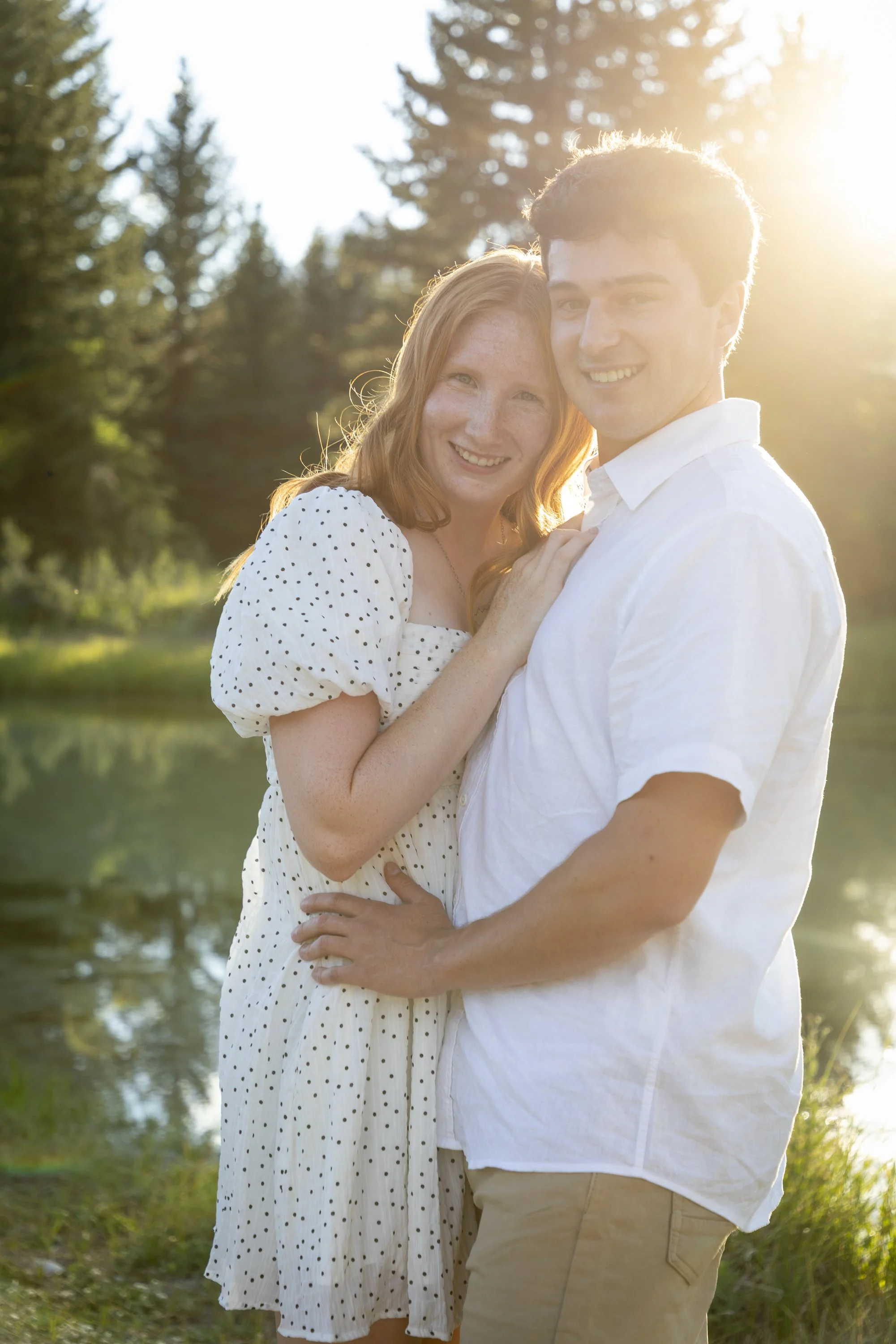 A smiling couple, a woman with red hair and freckles wearing a white dress with black polka dots, and a man with dark hair wearing a white shirt and beige trousers, standing outdoors near a body of water during sunset or sunrise.