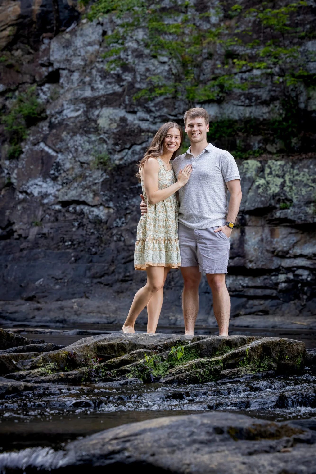 A smiling young couple stands on rocks beside a river with a rocky cliff background, both appearing relaxed and happy.