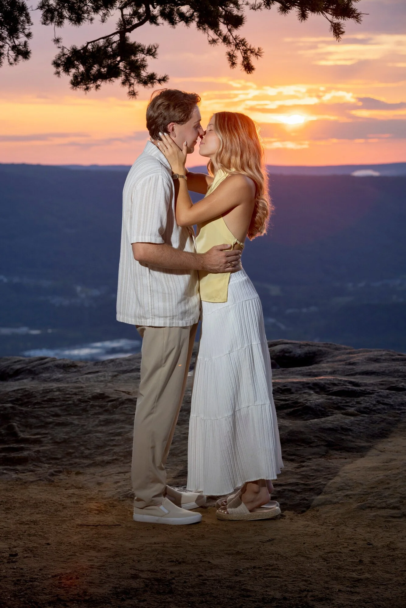 A couple is standing close together, touching noses and holding each other against a sunset backdrop over water and mountains, with tree branches overhead.