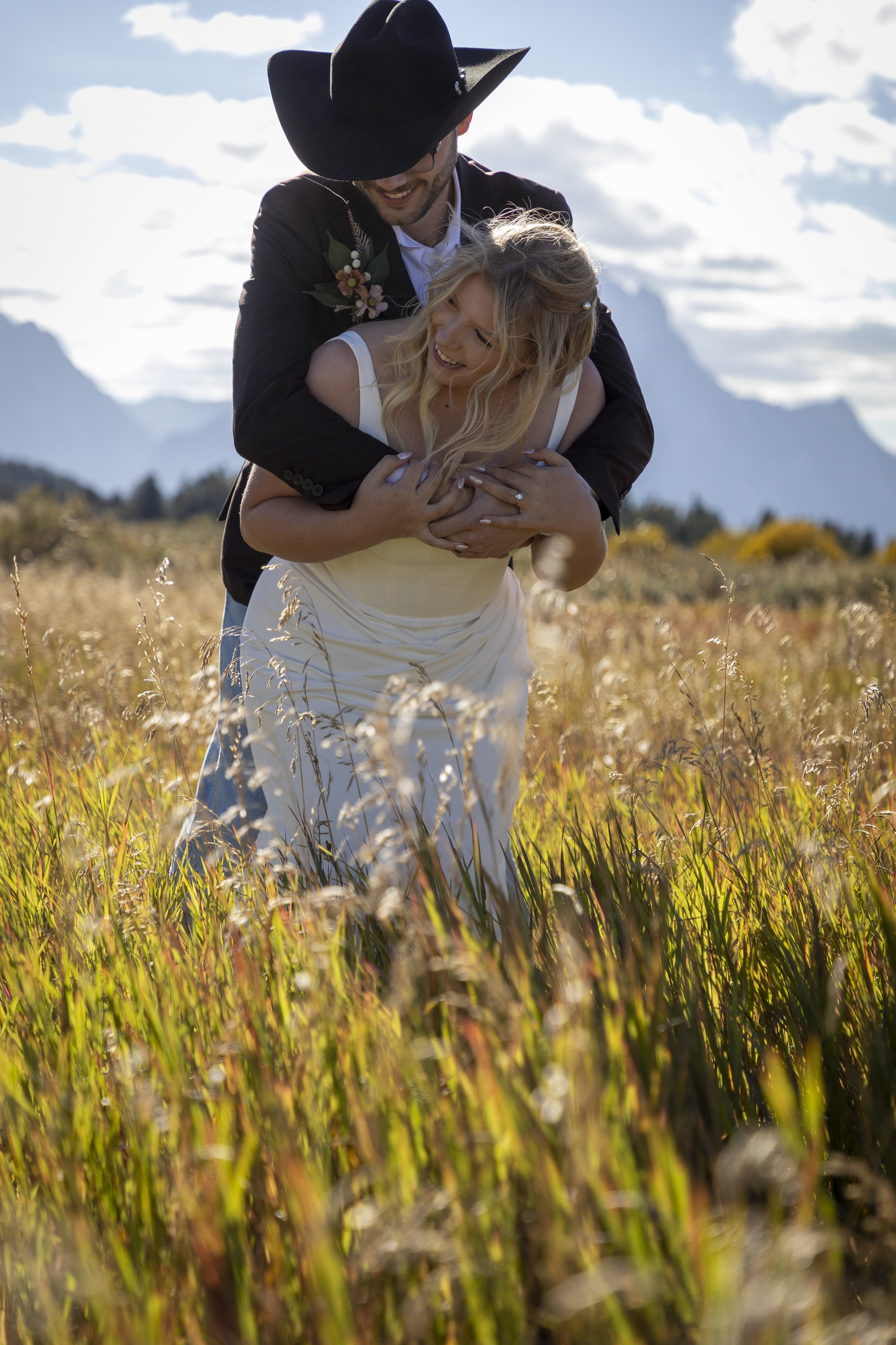A man in a black cowboy hat and a woman in a white dress are smiling and hugging in a field of tall grass with mountains and a partly cloudy sky in the background.