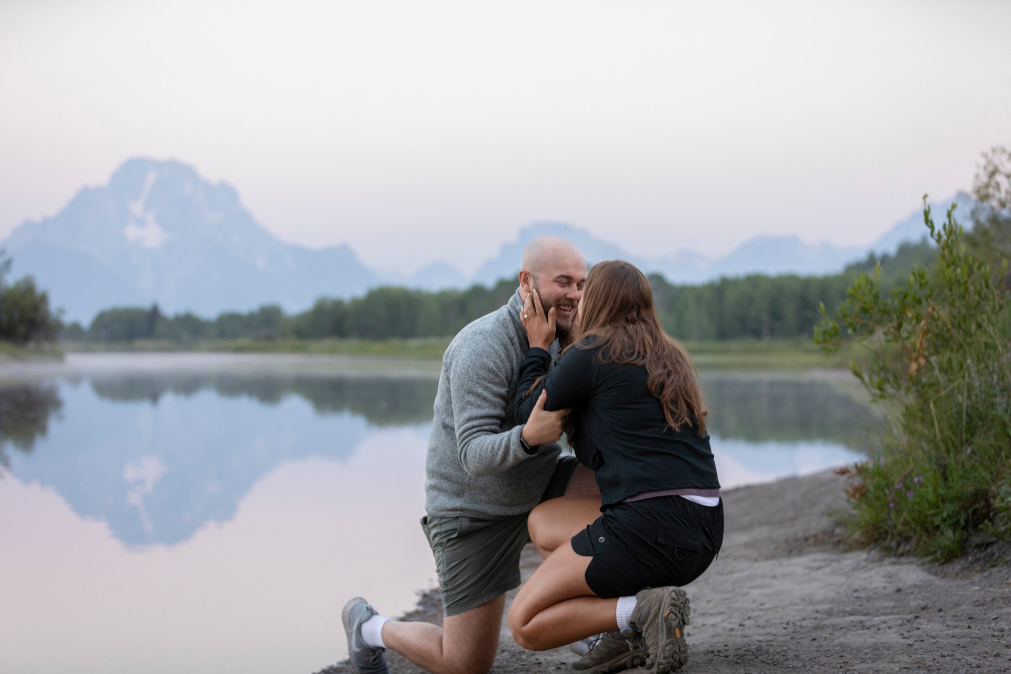 A man and woman are sharing a romantic moment by a lake with mountains in the background. The man is kneeling and the woman is embracing him, both smiling and enjoying each other's company in an outdoor setting.