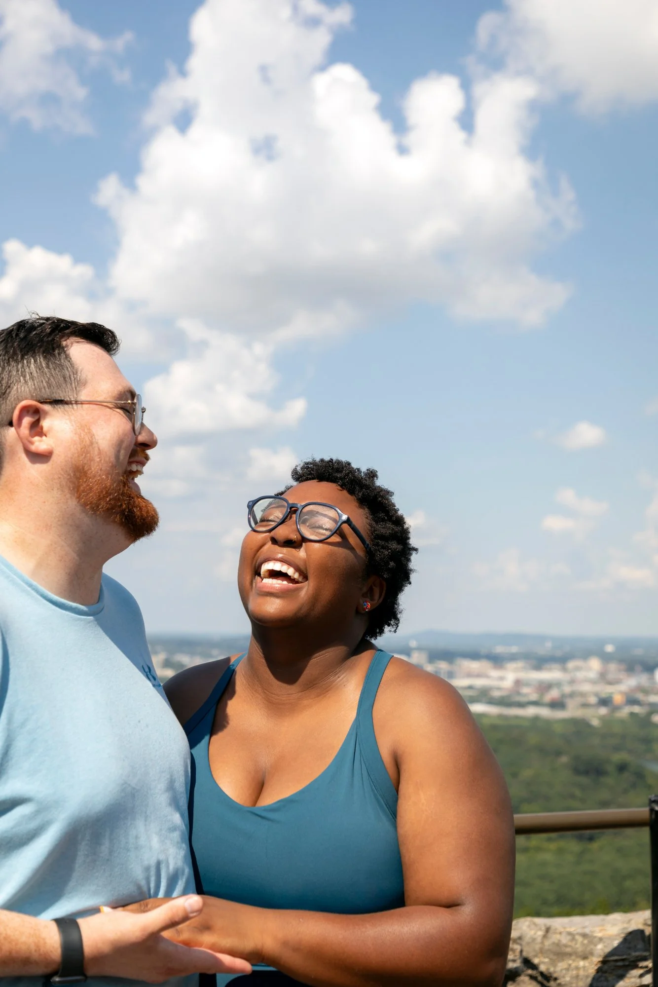 Two people smiling and looking at each other outdoors against a cloudy sky, with a city skyline in the background.