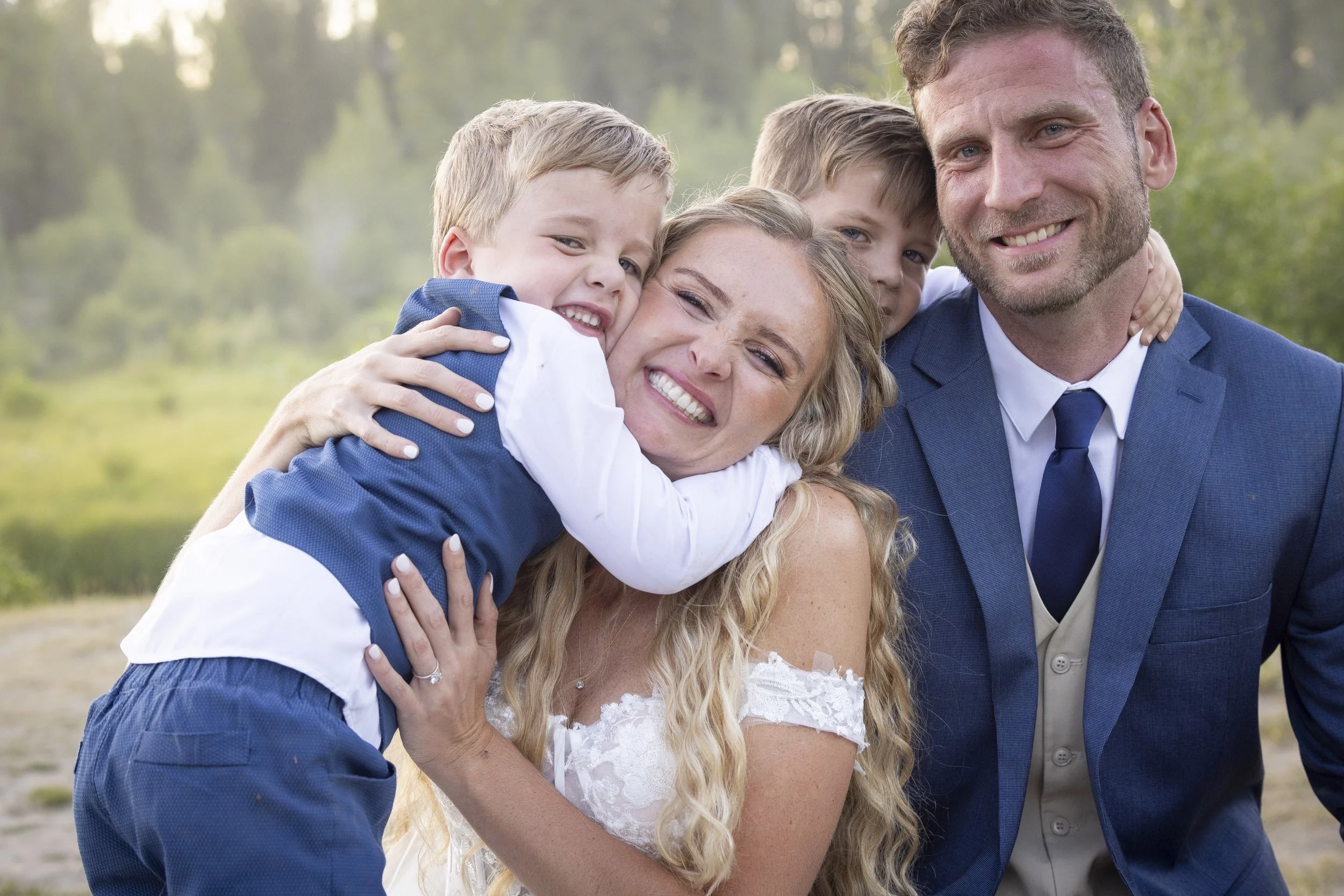 A family of five smiling and hugging outdoors on a sunny day with trees in the background. The woman in the center is surrounded by two young boys and is hugging a young boy while another man stands beside them.
