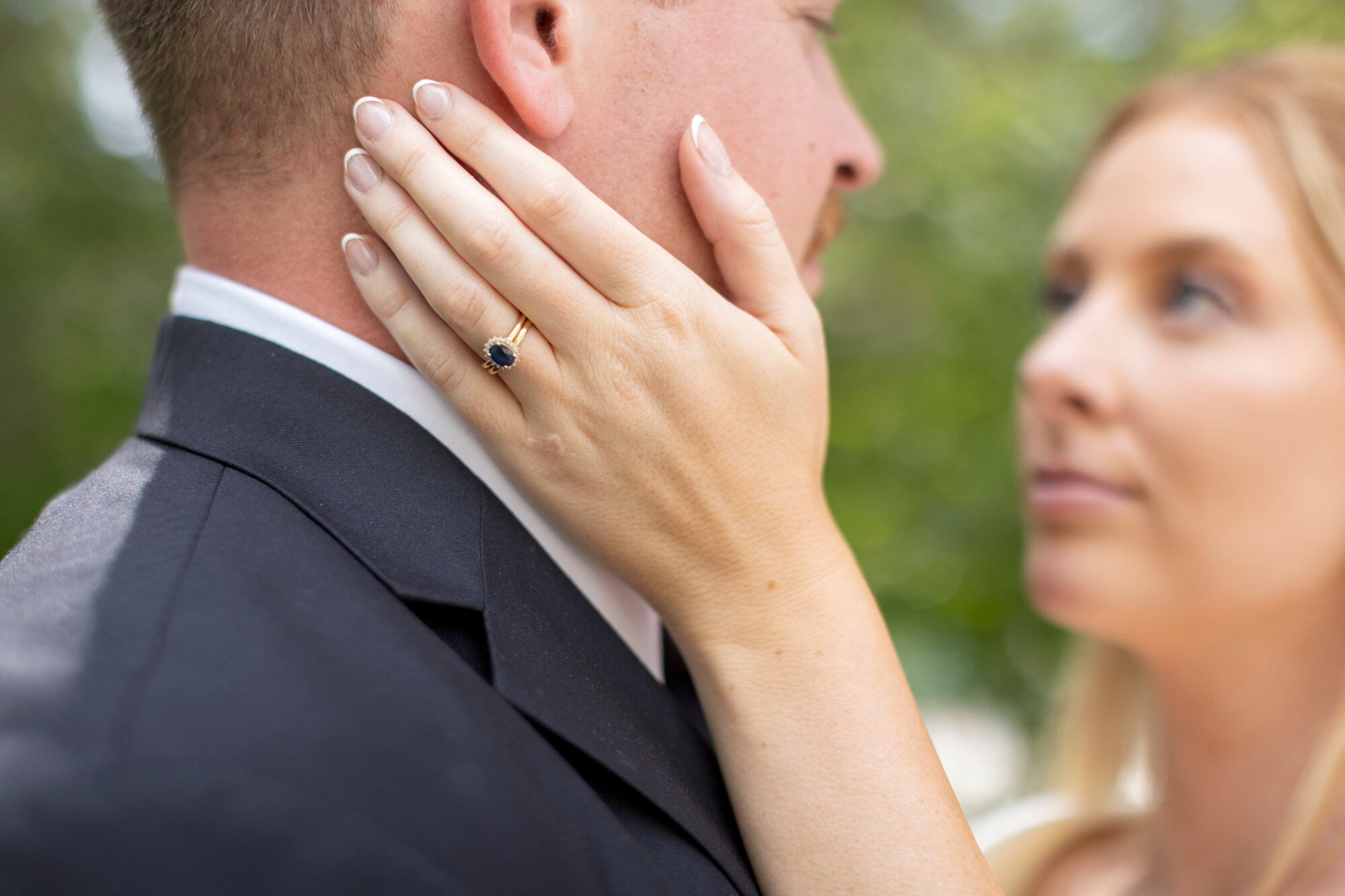 A woman with her hand on a man's face, showing a ring with a dark blue stone on her finger, in an outdoor setting with blurred greenery in the background.