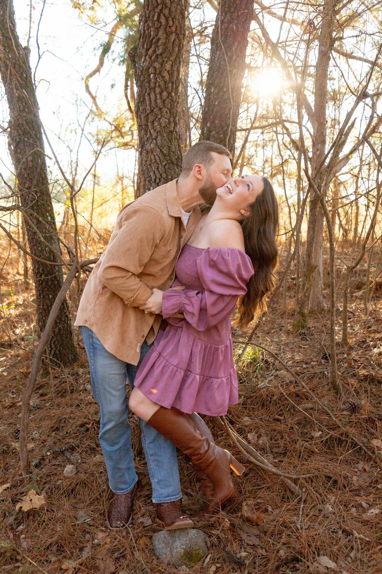 A couple sharing a romantic moment in a wooded area during fall, with sunlight filtering through the trees. The man is kissing the woman on the cheek, and they are smiling happily.
