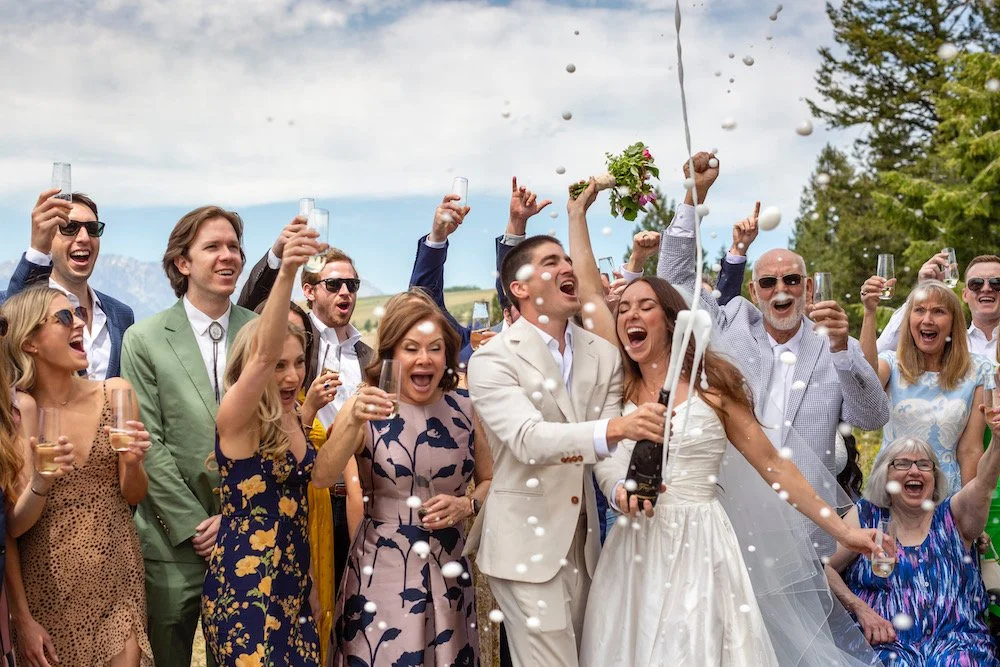 People celebrating outdoors, dressed in formal and semi-formal attire, with some wearing sunglasses, holding champagne glasses, and cheering as a bride and groom open a champagne bottle with corks and foam flying.