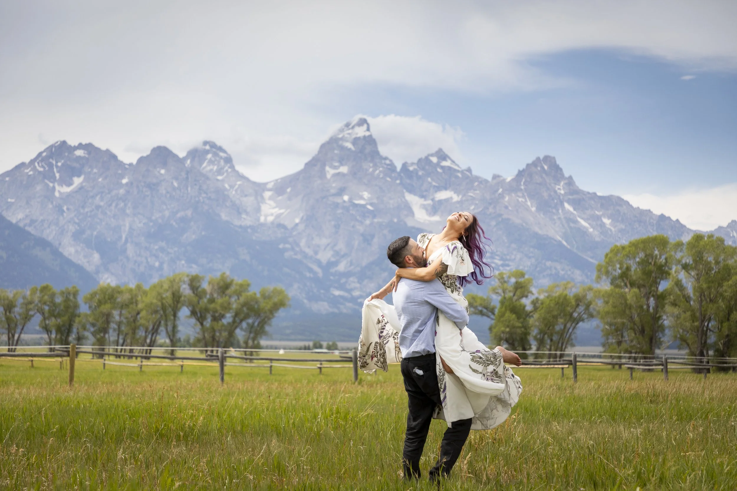 A couple, with the man lifting the woman, standing in a grassy field with trees and a wooden fence, mountains in the background, blue sky with some clouds.