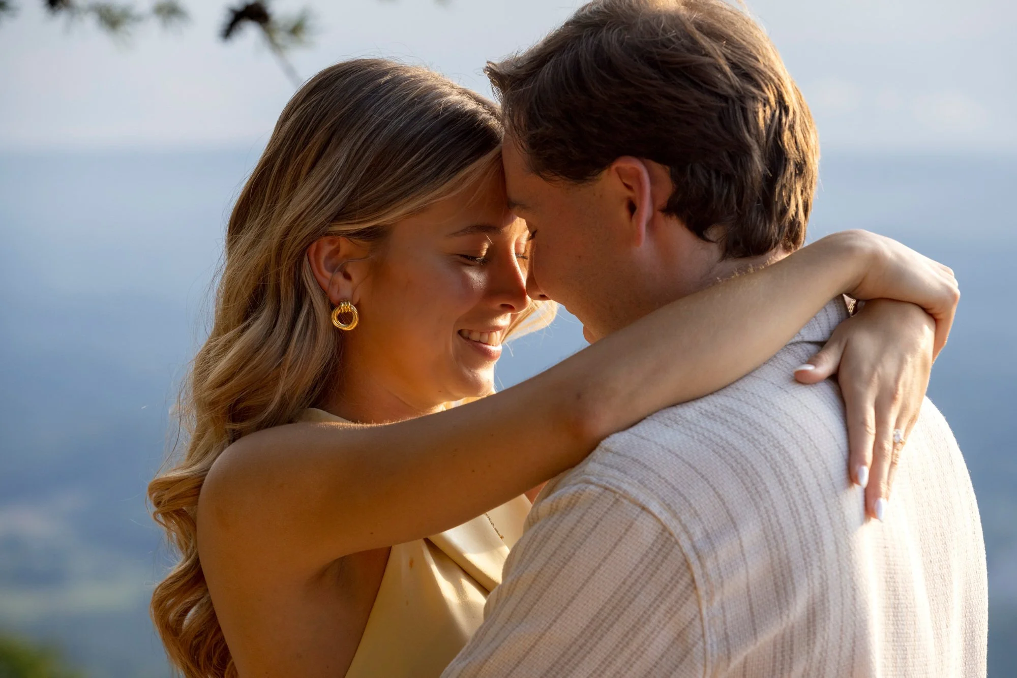 A couple embracing outdoors with their foreheads touching, eyes closed, smiling, against a background of blue sky and tree branches.