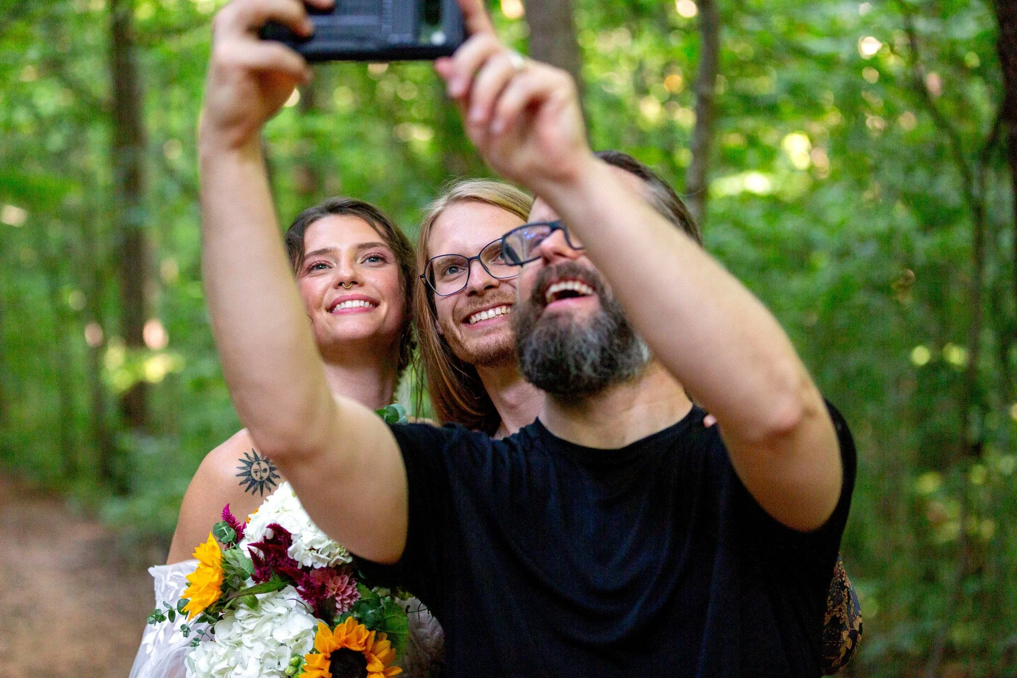 Three people taking a selfie in the woods. The person in the foreground is a man with glasses and a beard, smiling. Behind him is a woman with long hair, also smiling, holding a bouquet of flowers. Behind her is another woman with long hair, smiling.