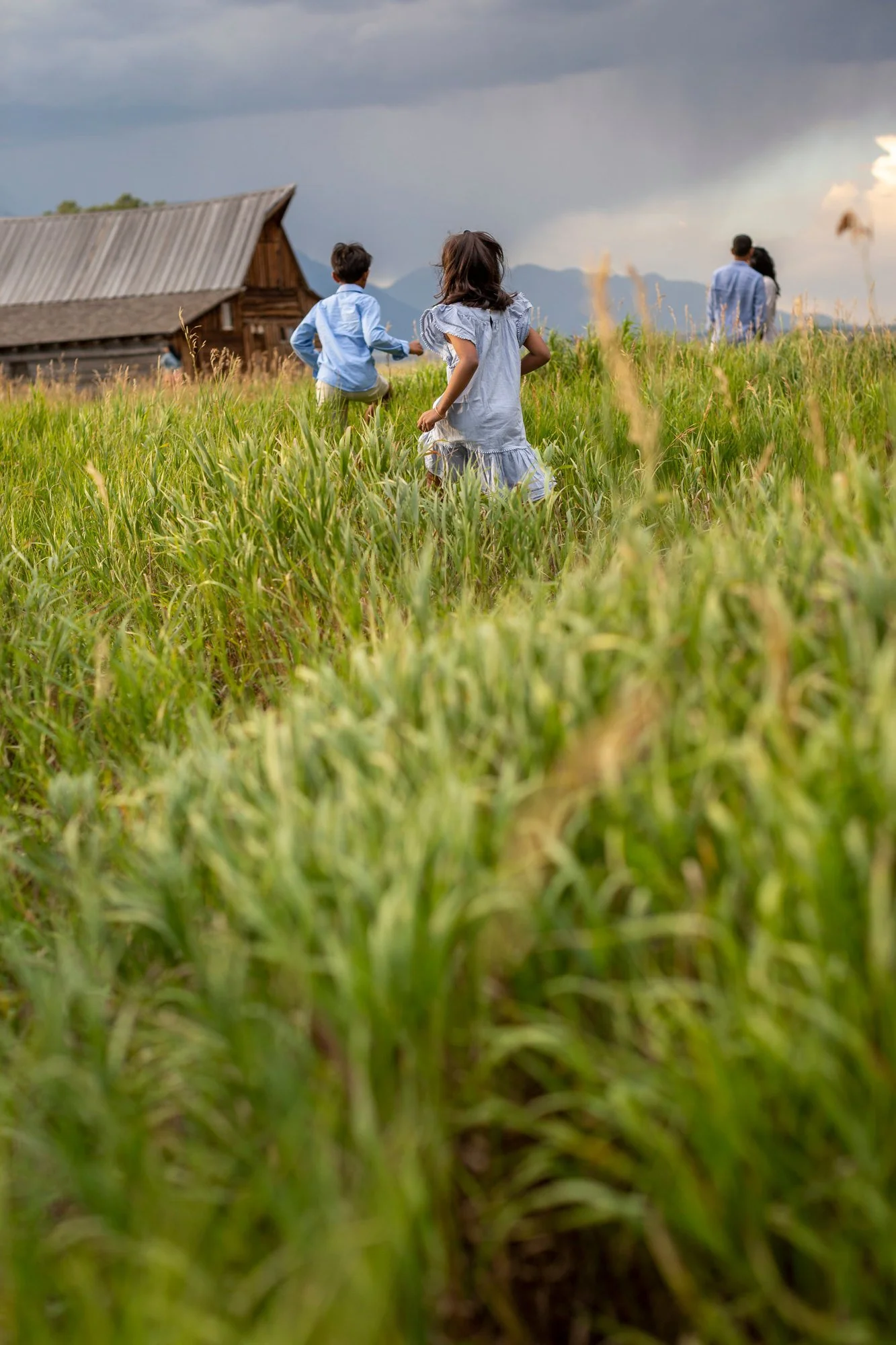 Children and adults walking through a grassy field with a wooden barn in the background and mountains under a cloudy sky.