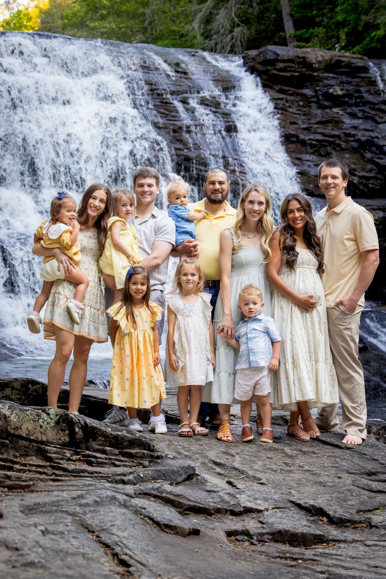A multigenerational family of thirteen people, including children and adults, standing together on rocks near a waterfall during daytime, with greenery in the background.