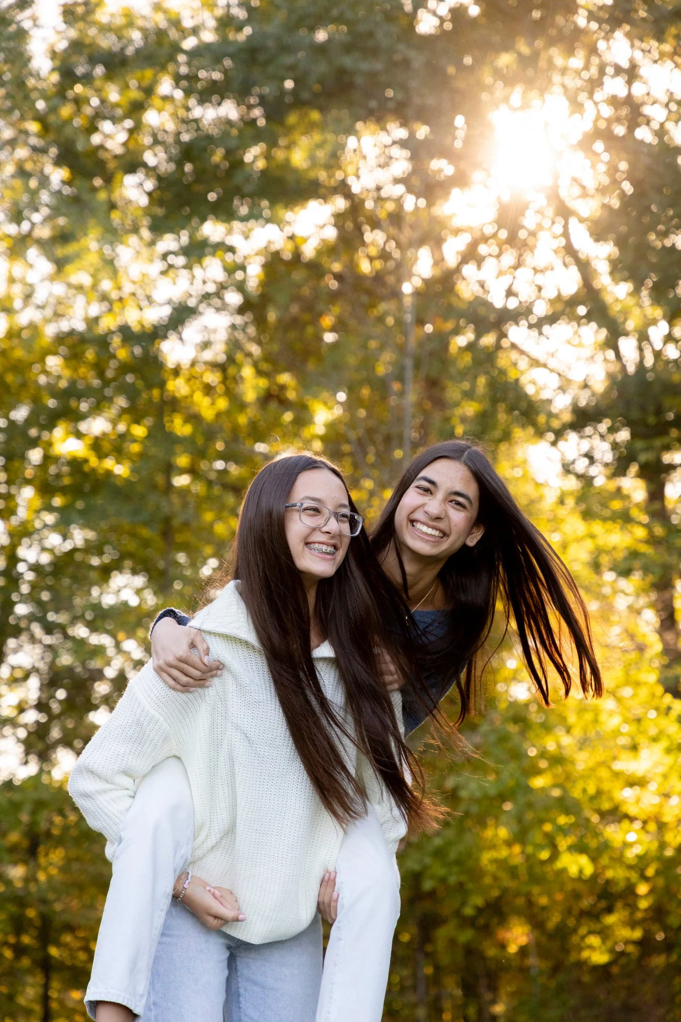 Two young women smiling and hugging outdoors with trees and sunlight in the background.