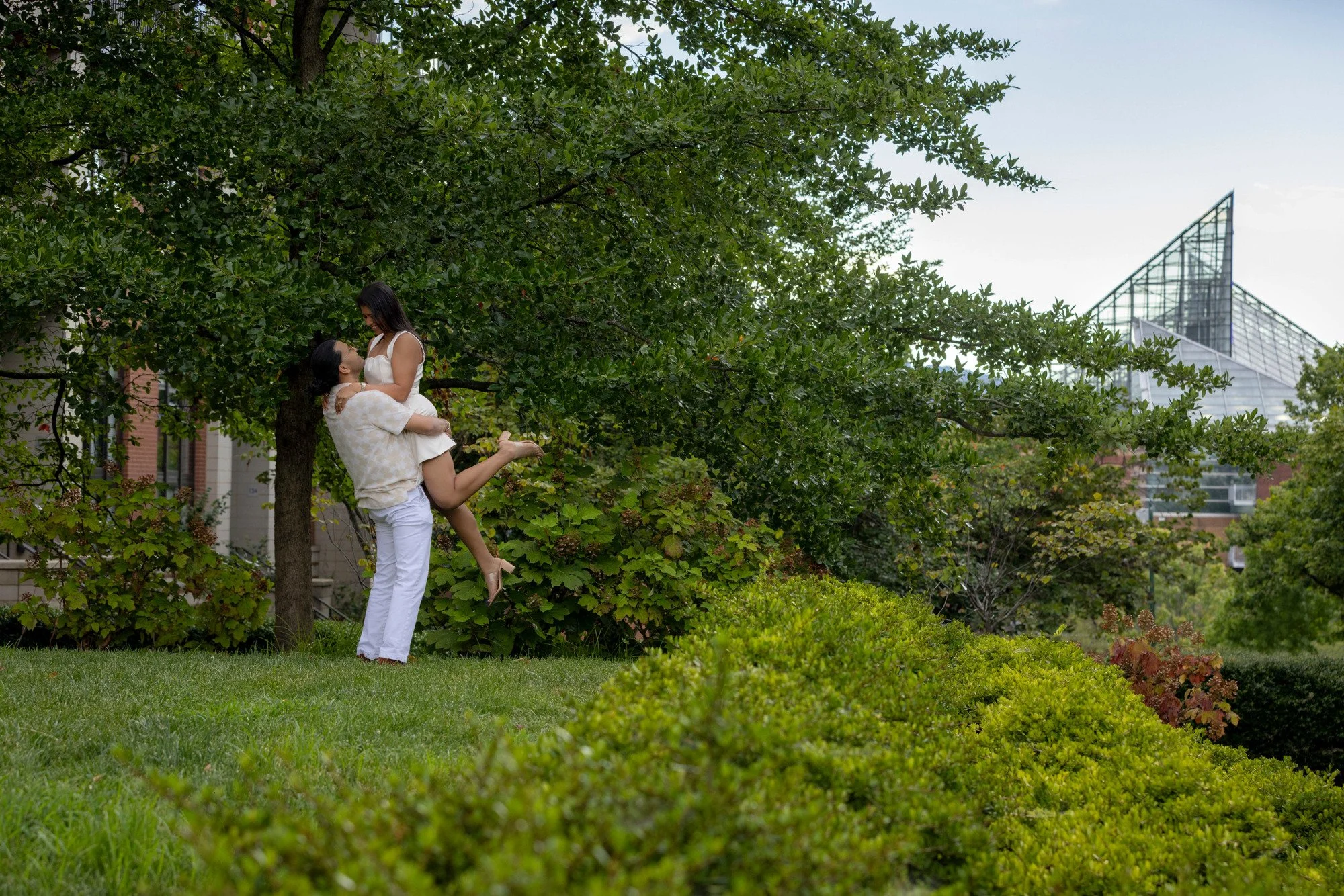 Two women share an affectionate moment in a park, with one woman lifting the other in her arms under a large tree, surrounded by green foliage and city buildings in the background.