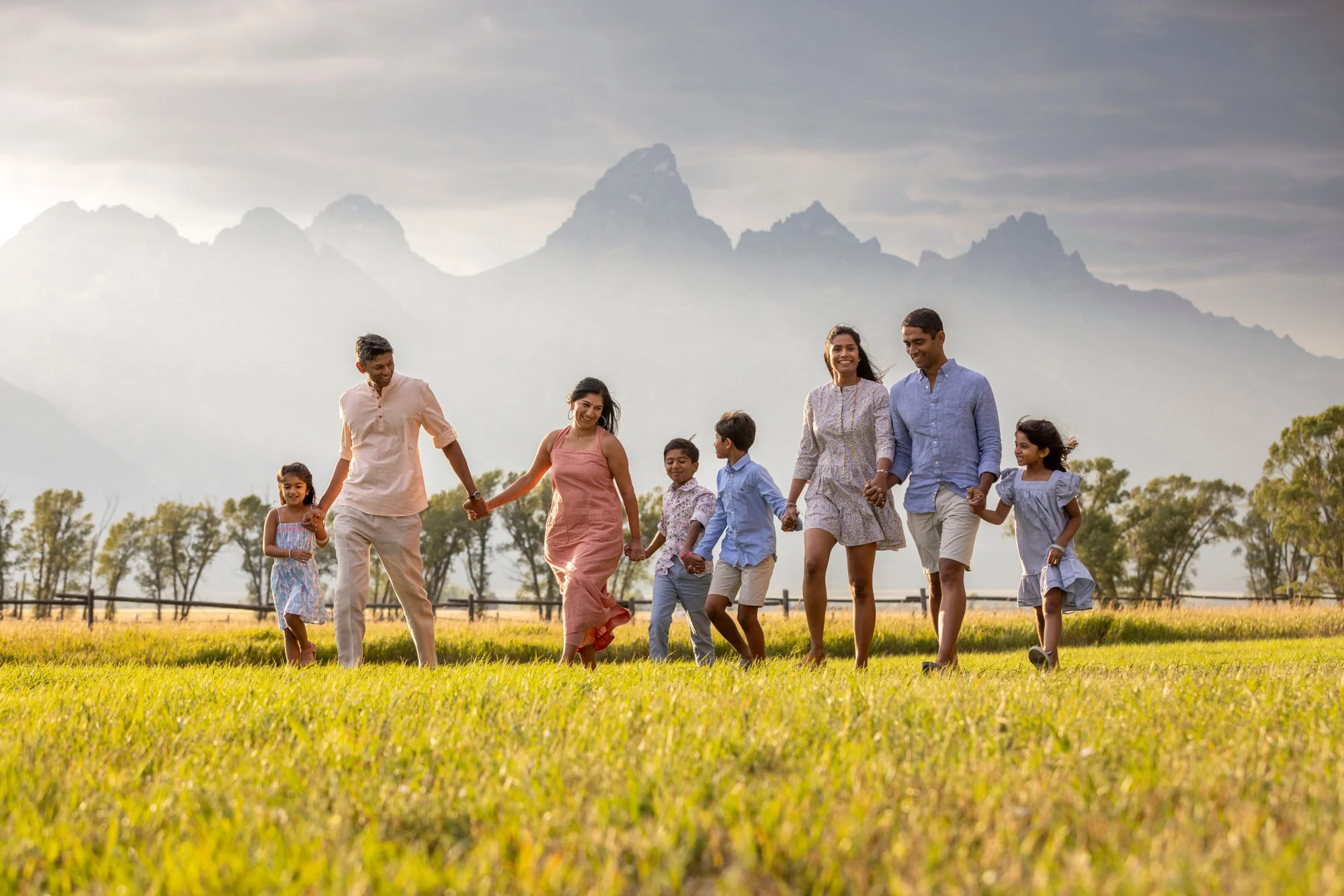 A family of nine people, including children, walking hand in hand across a grassy field with mountains in the background under a cloudy sky.