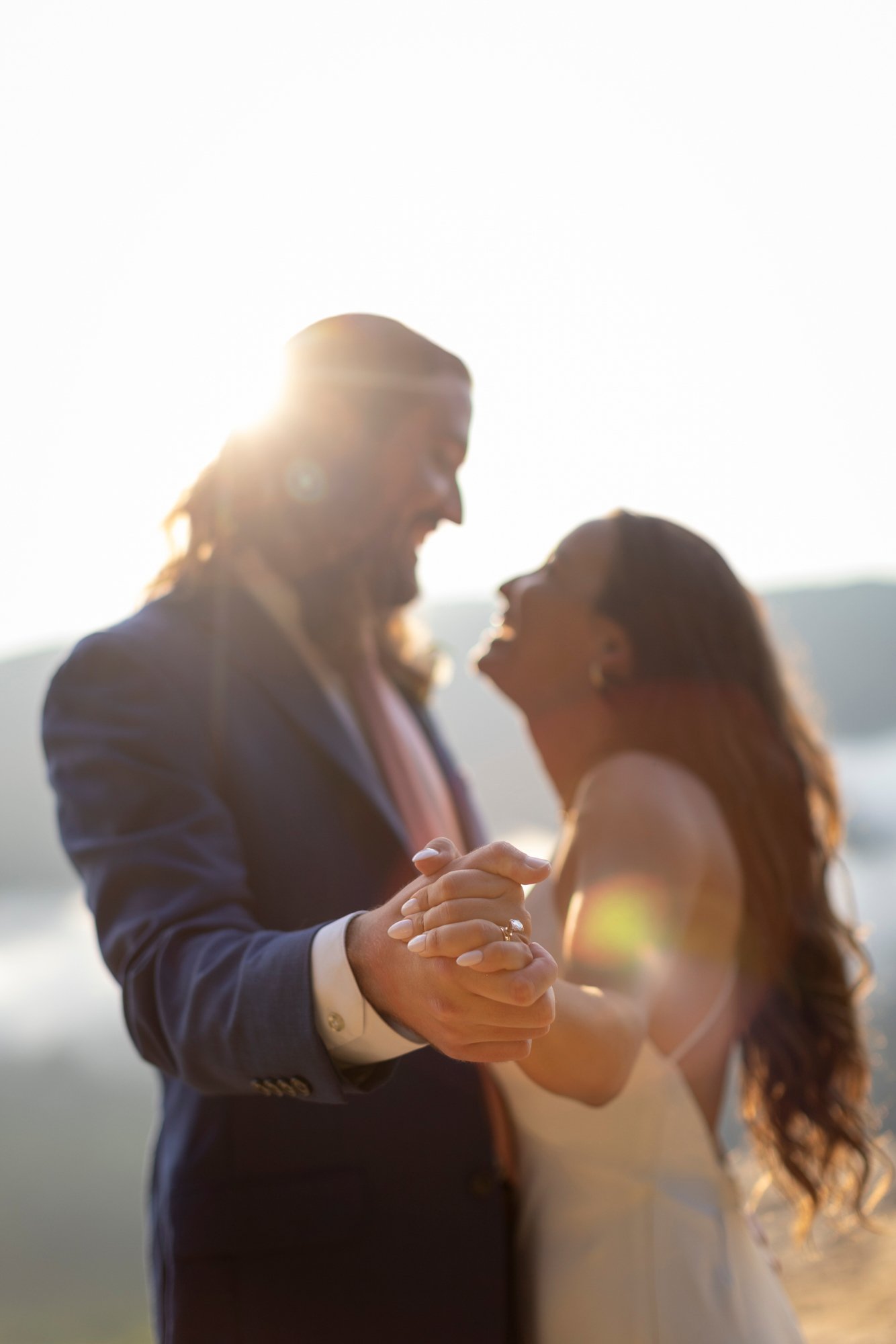 A couple holding hands and dancing outside during sunset, with the man wearing a dark suit and the woman in a wedding dress, smiling and looking at each other.
