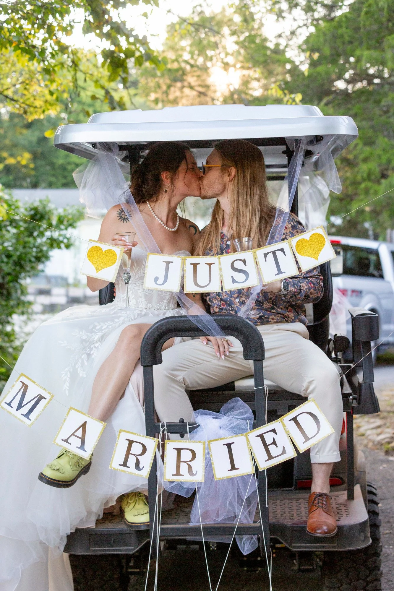 Two people kissing in wedding attire, sitting in a golf cart decorated with wedding signs, greenery in the background, sunset, one person wearing a wedding dress and pearl necklace, the other in patterned shirt and sunglasses.
