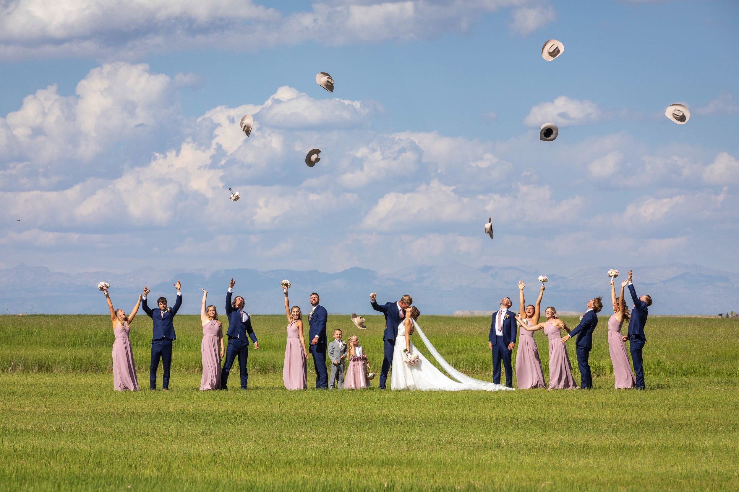 Group of wedding guests, including bridesmaids and groomsmen, celebrating outdoors in a field with a blue sky and clouds, as their hats are being tossed into the air.