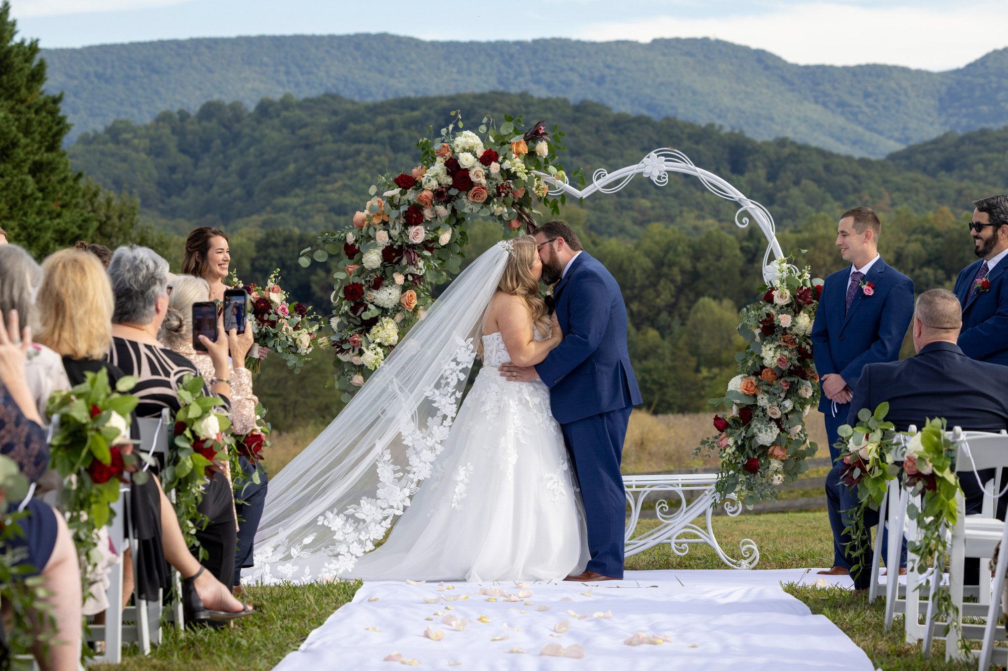 A wedding ceremony outdoors with a bride and groom kissing under a floral arch, surrounded by guests and scenic mountain landscape.