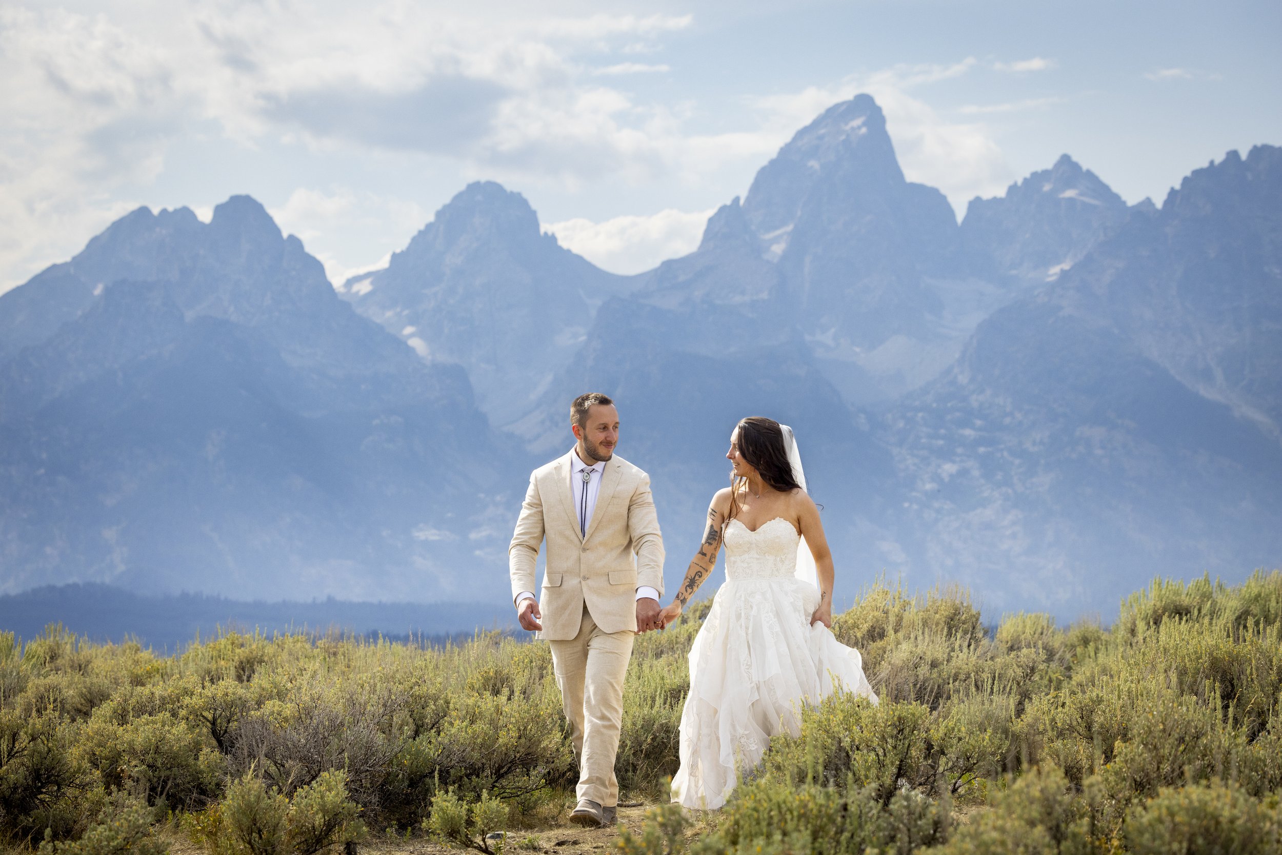A groom and bride walking hand in hand in a field with mountains in the background on a partly cloudy day.