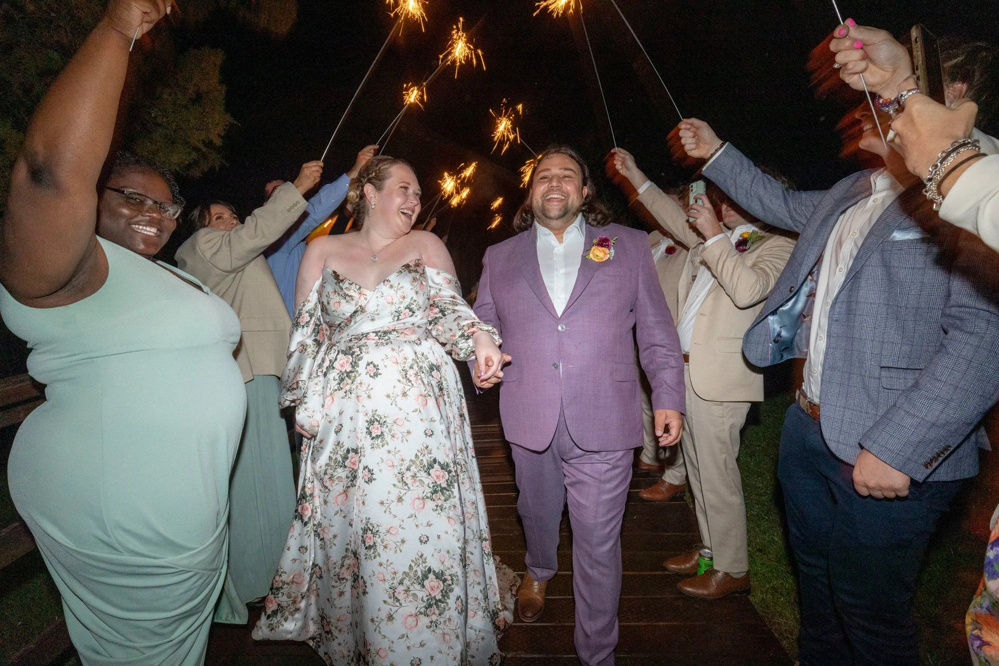 A joyous wedding celebration at night features a bride and groom holding hands and smiling, surrounded by friends holding sparklers in the air. The bride is wearing a floral dress, and the groom is in a purple suit with a boutonniere.