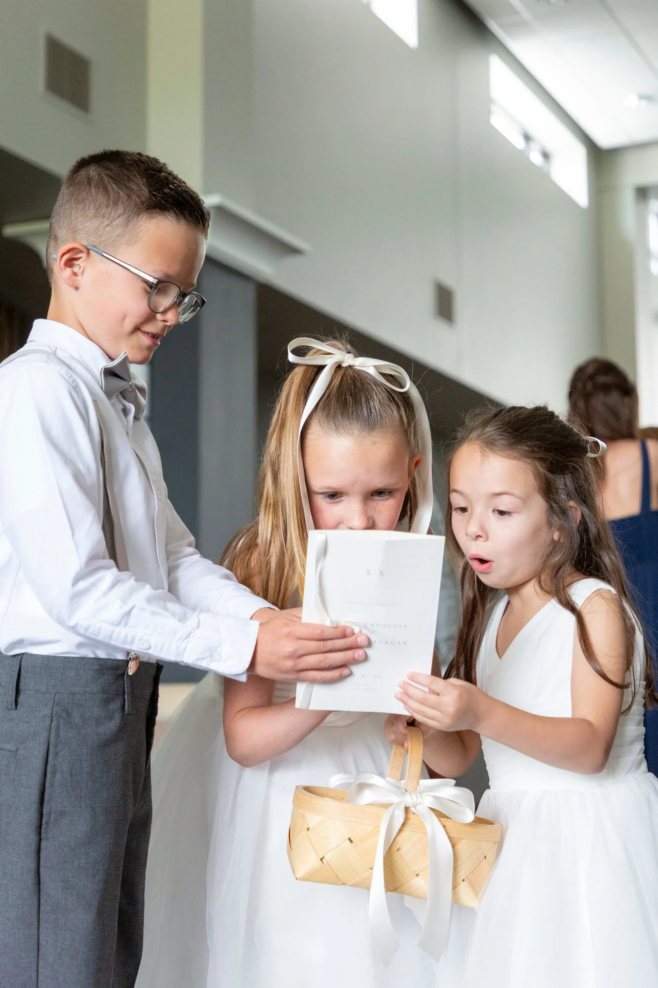 A young boy in glasses and formal attire showing a piece of paper to two young girls dressed in white at what appears to be a ceremony or celebration. One girl is holding a basket, and the other is looking at the paper with interest.