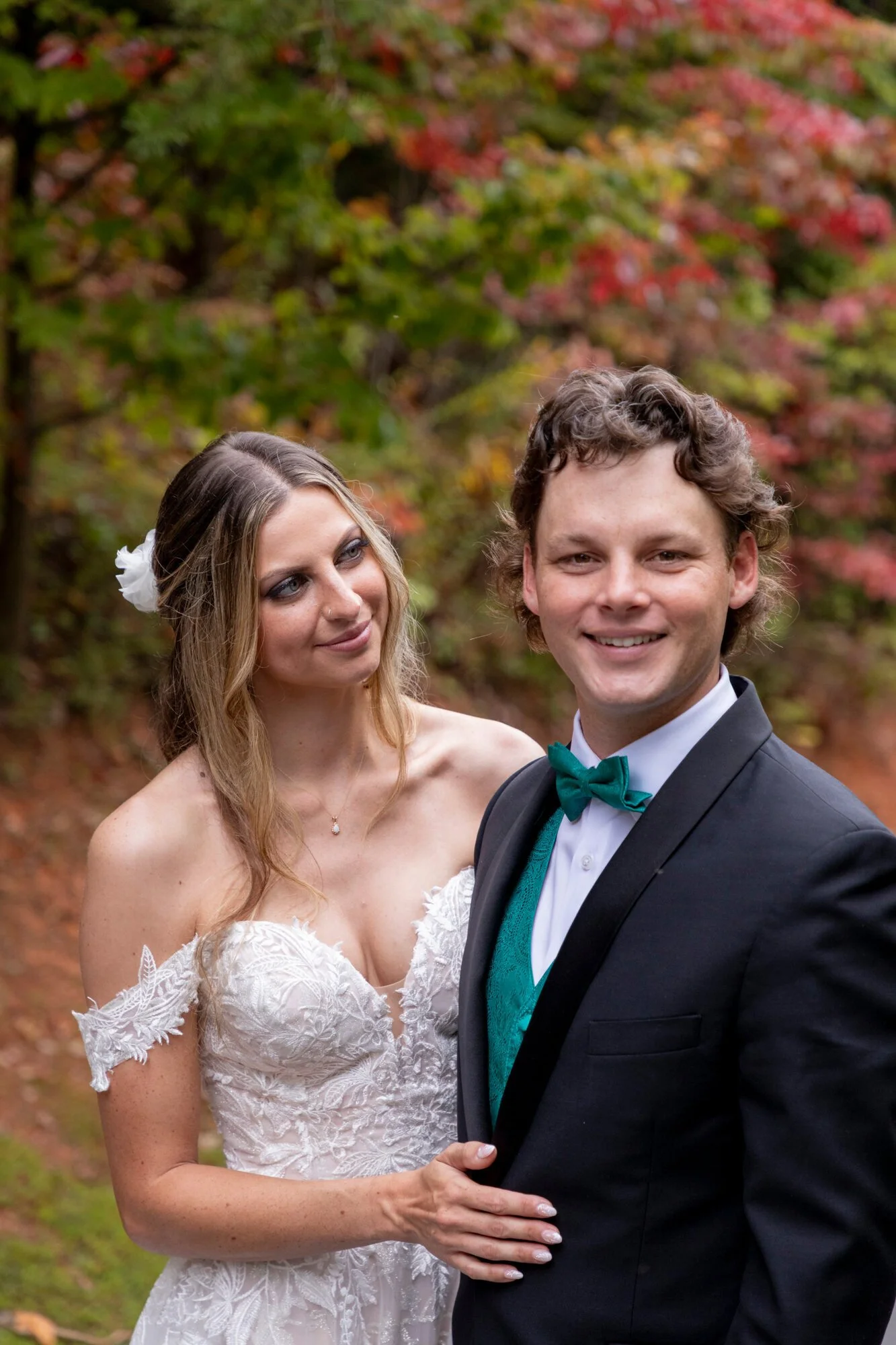 A smiling bride and groom standing outdoors among autumn foliage, with the bride wearing a white lace wedding dress and the groom in a dark tuxedo with a teal bow tie.