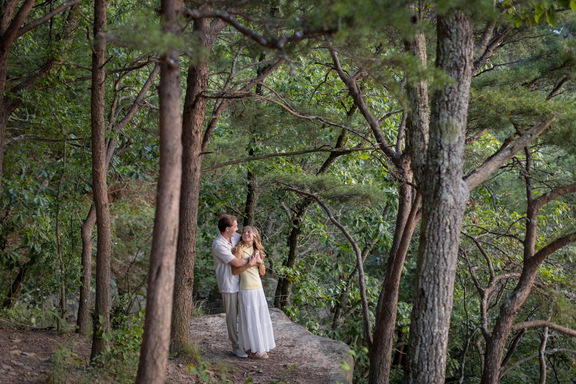 A couple standing on a path in a dense forest, embracing and gazing at each other, surrounded by tall trees and lush green foliage.