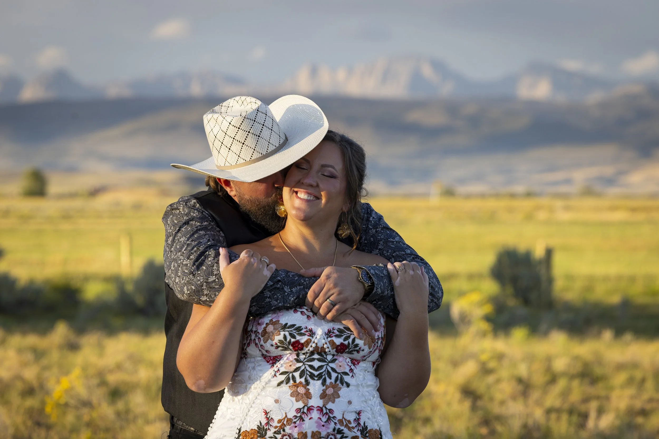 A couple embracing outdoors, with the man kissing the woman on the cheek as she smiles with her eyes closed, in a field with a mountain range in the background.