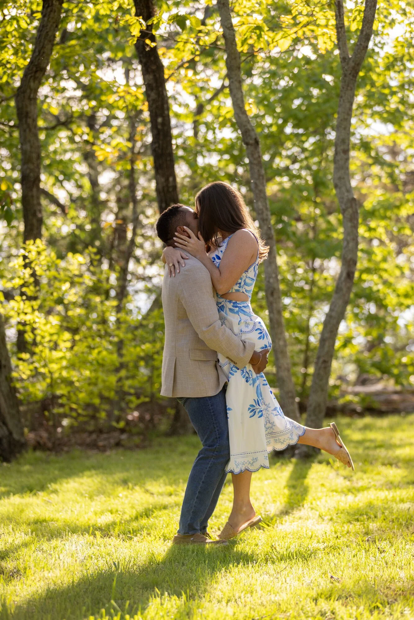 A couple kissing outdoors in a park, with the man lifting the woman, surrounded by green trees and sunlight.