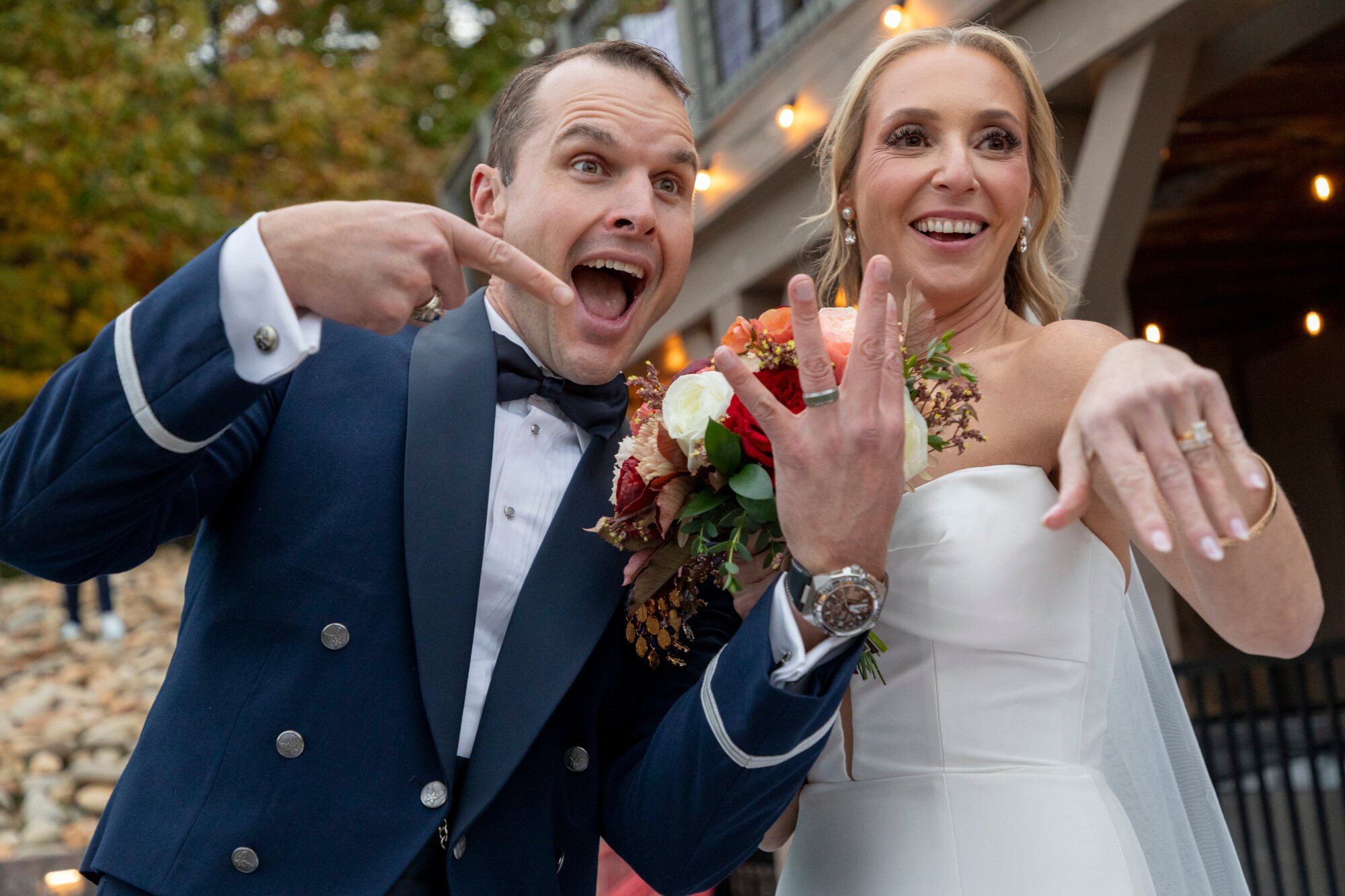 A smiling couple, standing outdoors at sunset, making playful gestures toward the camera. The man is in a dark blue tuxedo with silver buttons, and the woman in a white wedding dress holding a bouquet of flowers.