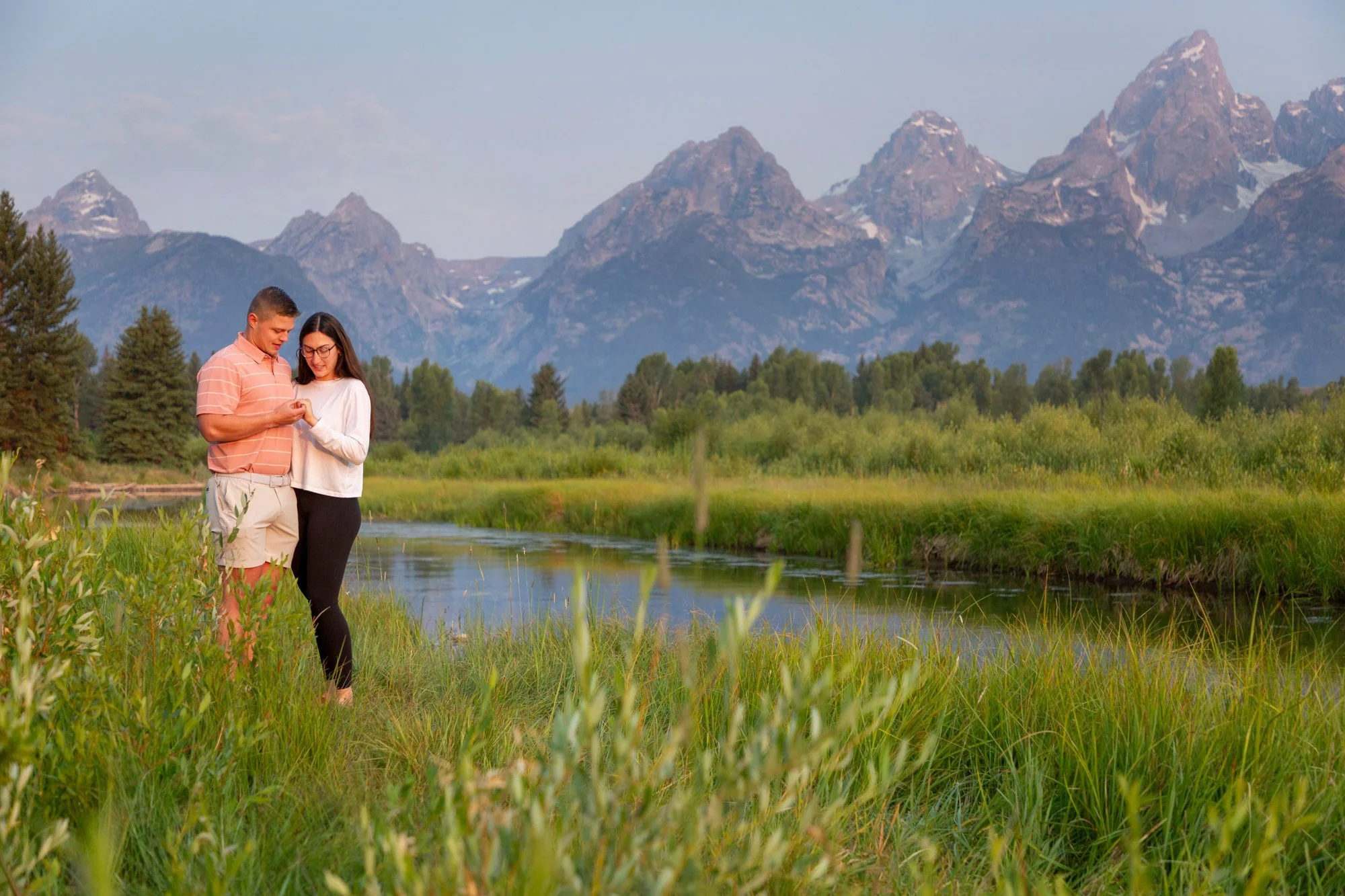 A young man and woman standing next to each other by a river, looking at a phone, with mountains in the background and green trees and grass surrounding them.