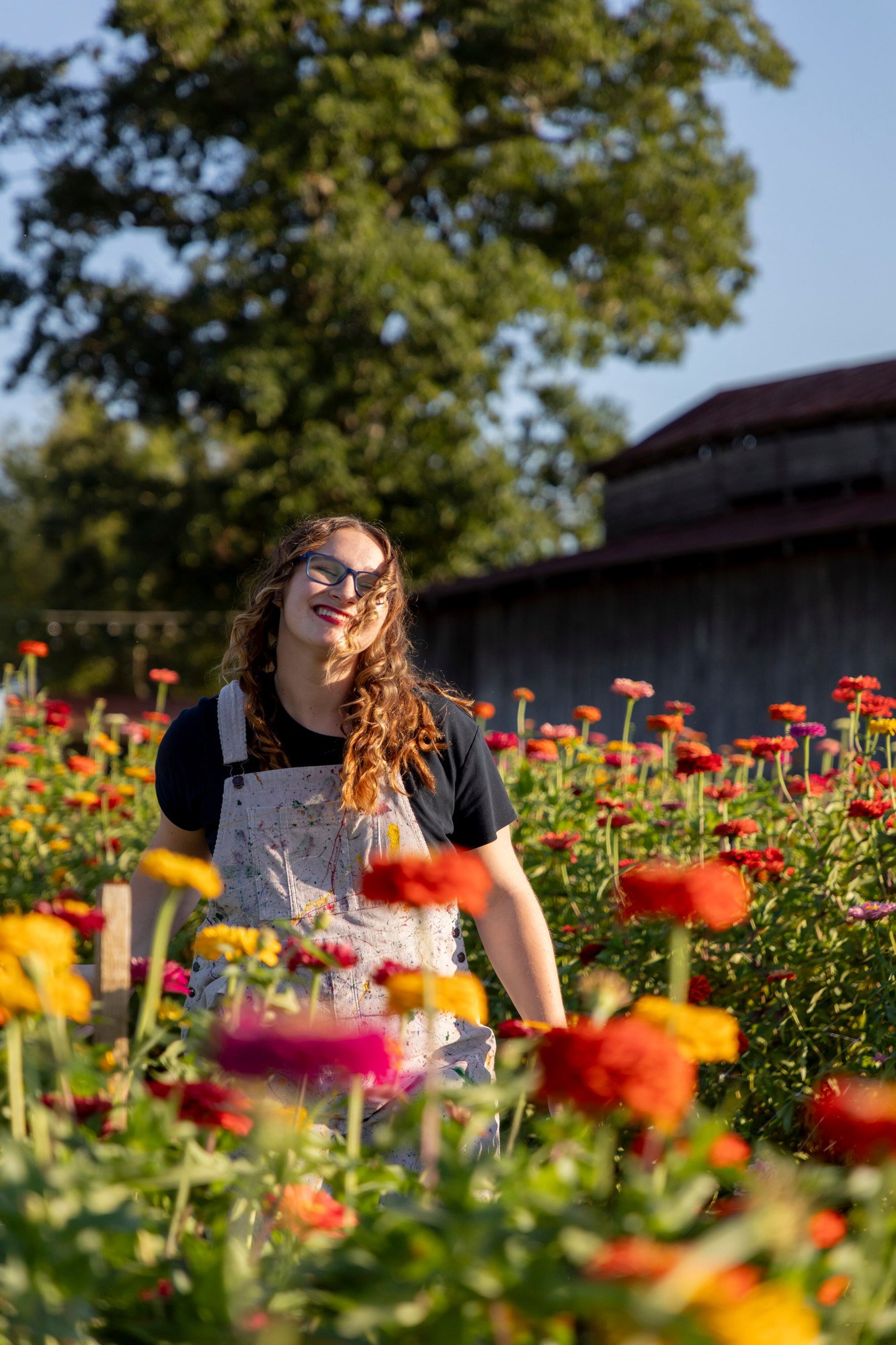 A woman with glasses and curly hair smiling with closed eyes in a flower garden during daytime, wearing a black shirt and a paint-splattered apron.