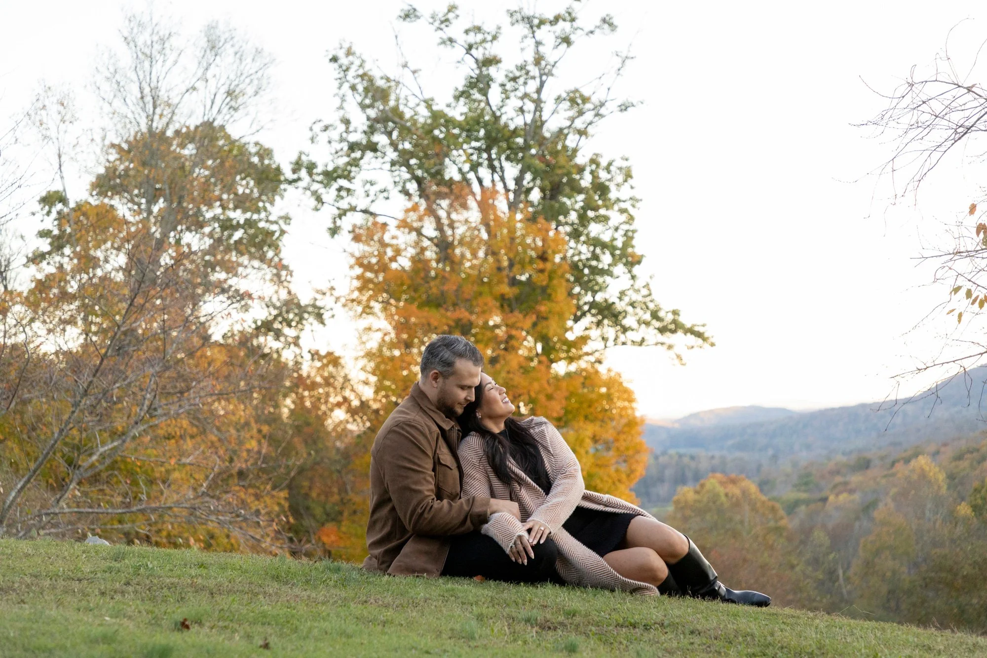 A couple sitting on the grass in a park during fall, embracing with trees displaying colorful autumn leaves in the background.