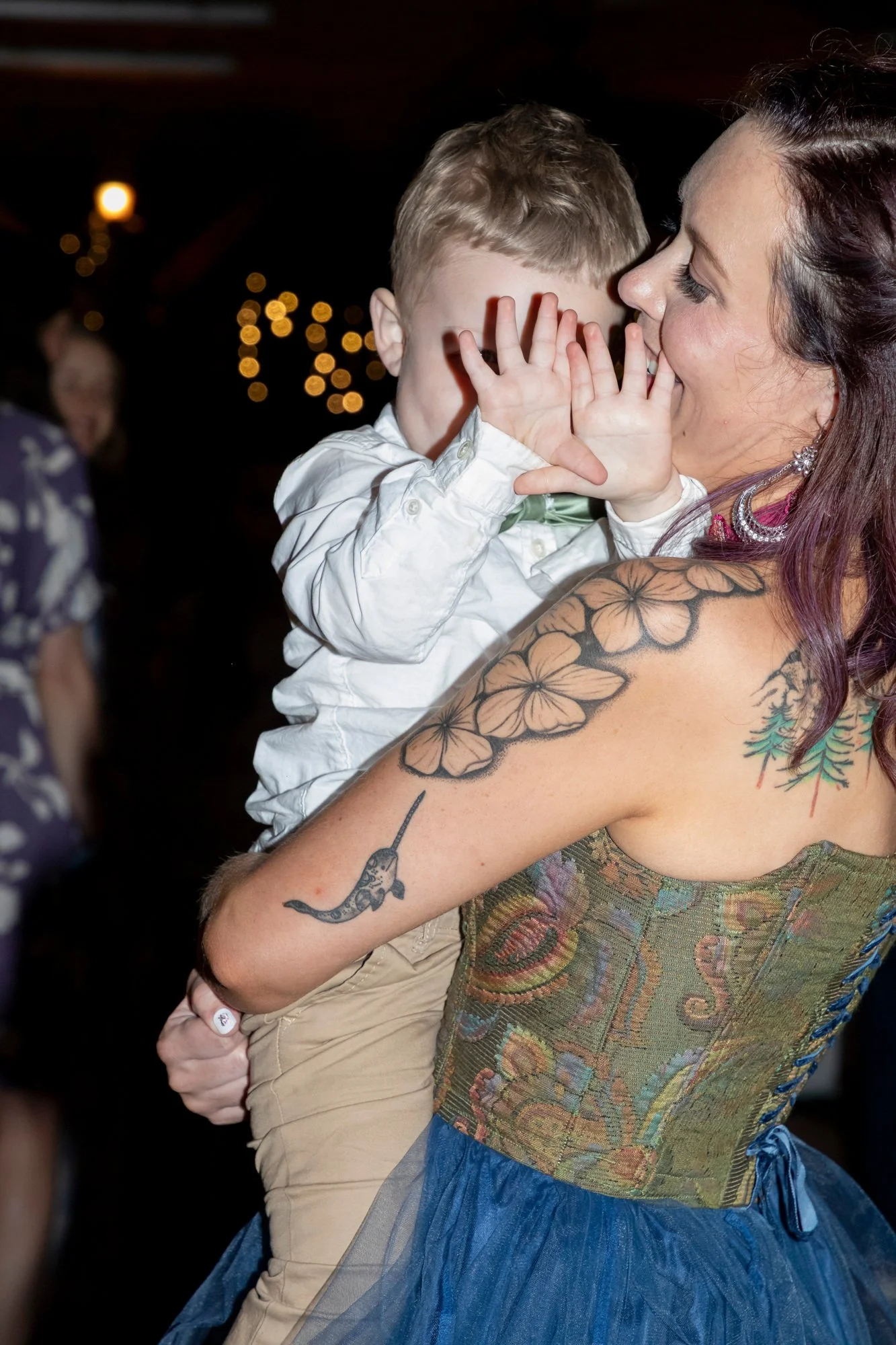A woman with tattoos on her arm holding a young boy close to her face, both smiling and laughing, in a dark setting with blurred lights in the background.