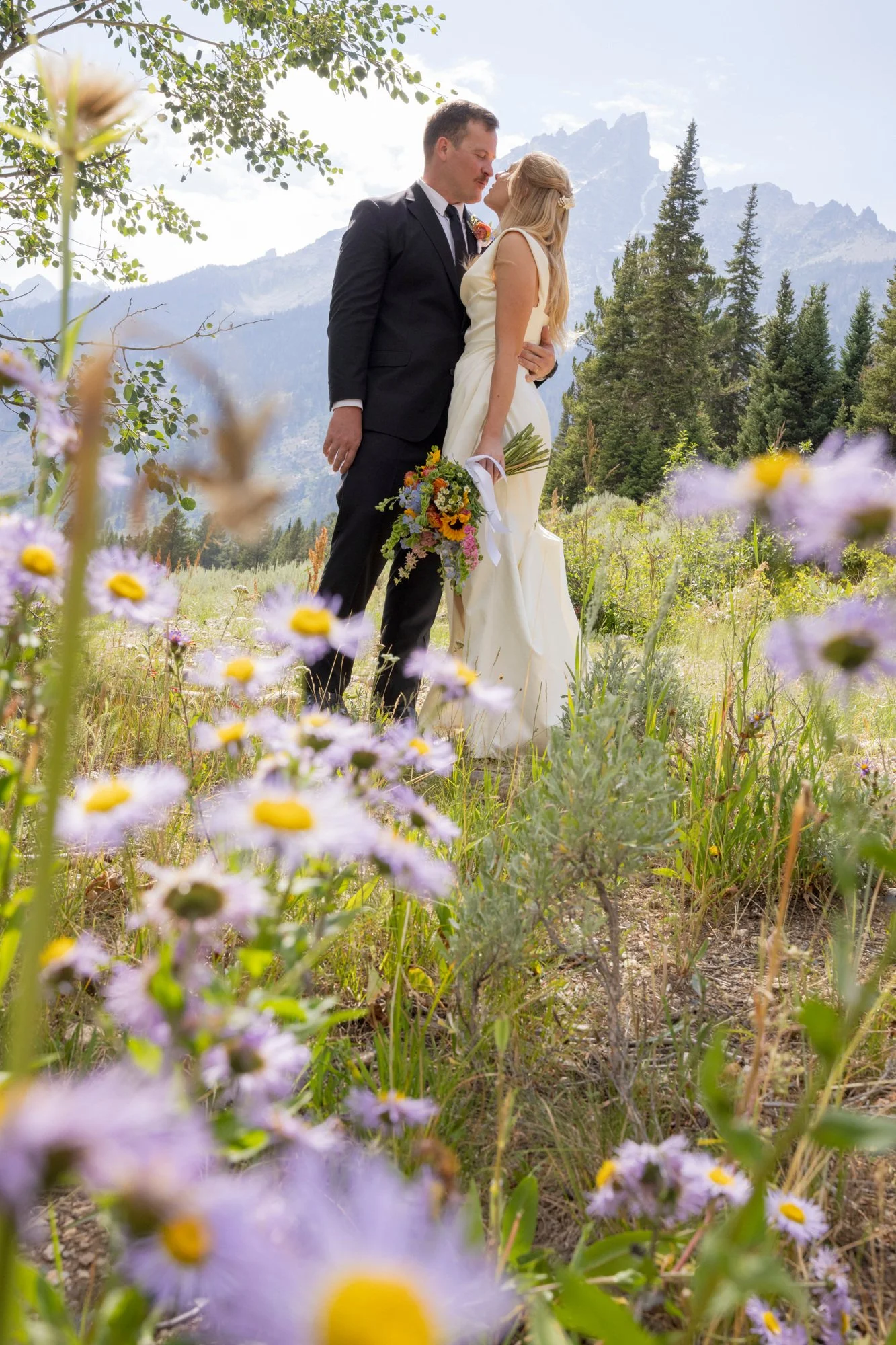 A couple dressed in formal attire, standing close together outdoors among wildflowers with mountain peaks and trees in the background.