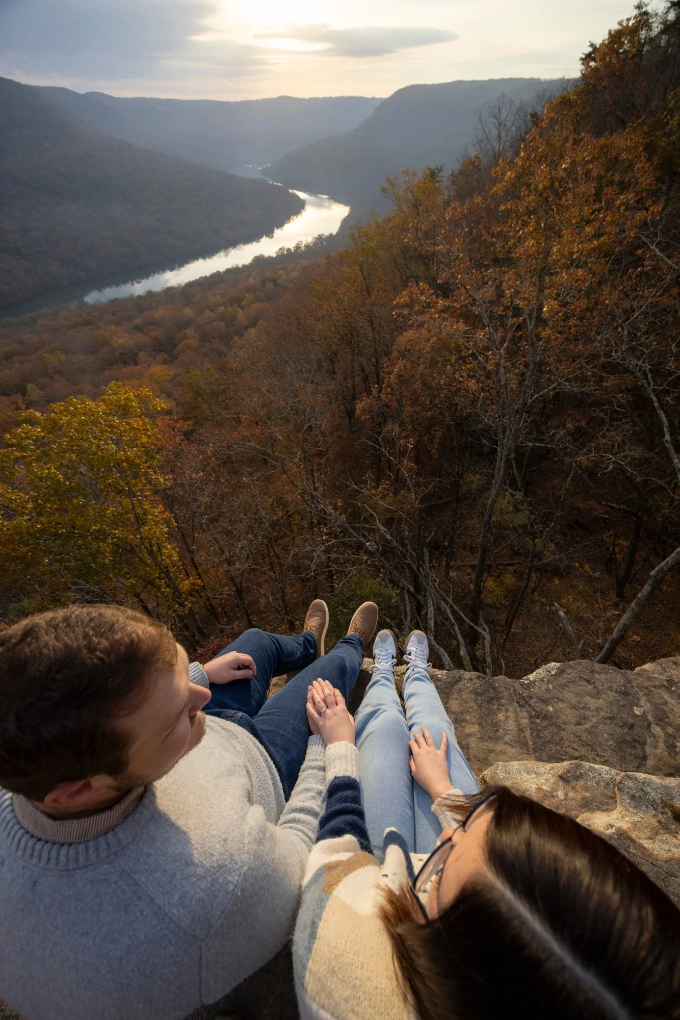 A group of people sitting on a rocky ledge overlooking a scenic valley with a river winding through it, surrounded by autumn-colored trees and mountains in the background.