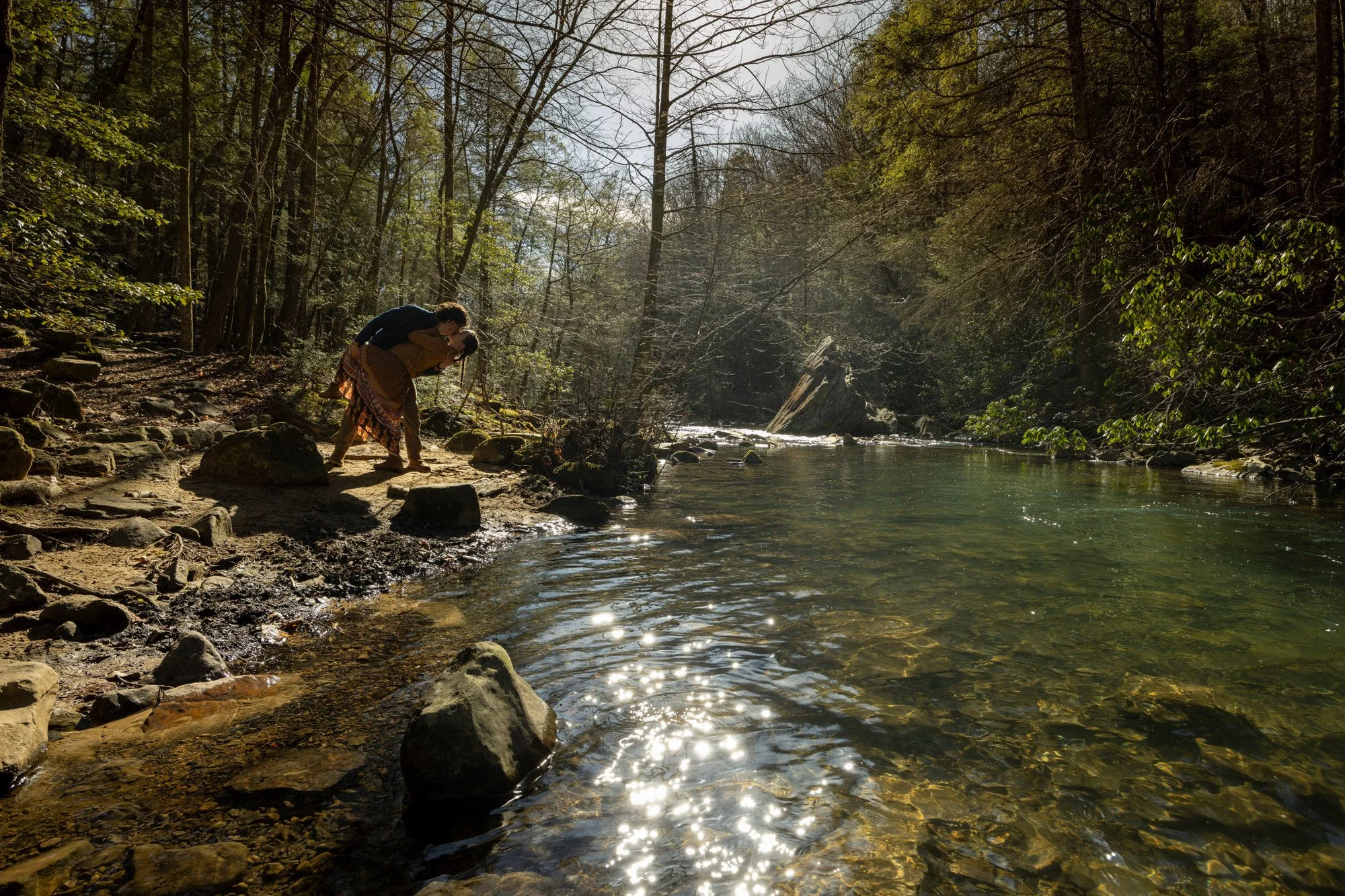 Two people are on a rocky shoreline next to a river in a dense forest. One person is bent over, leaning on a rock, while the other stands behind, leaning over and appears to be helping or supporting them. The sunlight filters through the trees, creat