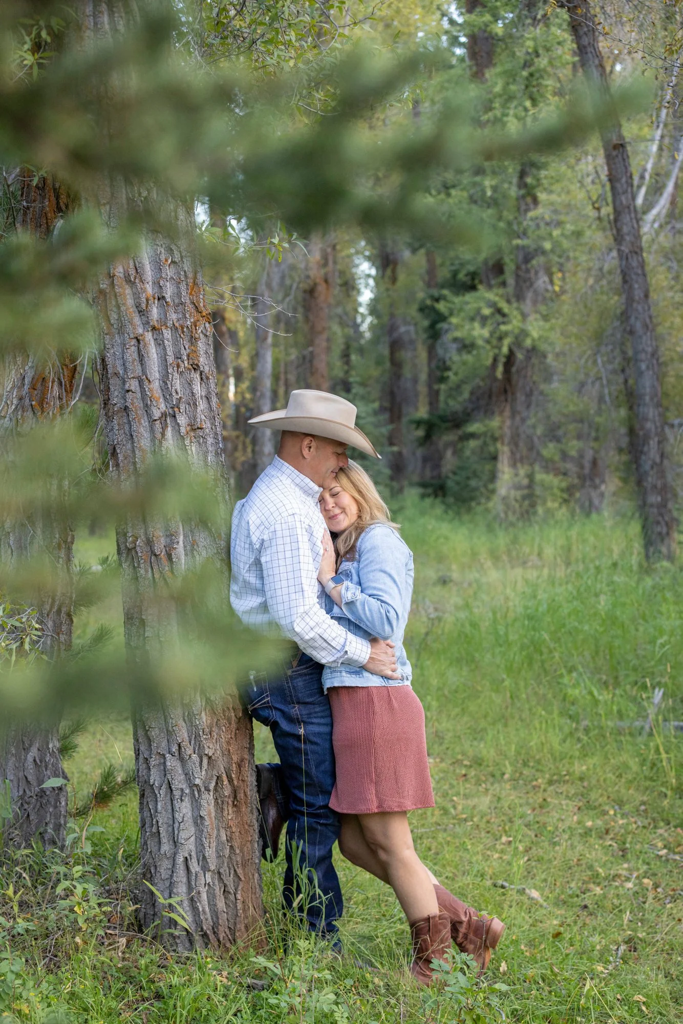 A couple embracing outdoors beside a tree in a forest, with lush green grass and trees in the background. The man is wearing a cowboy hat, a checkered shirt, and jeans, and the woman is in a denim jacket, pink skirt, and brown boots.