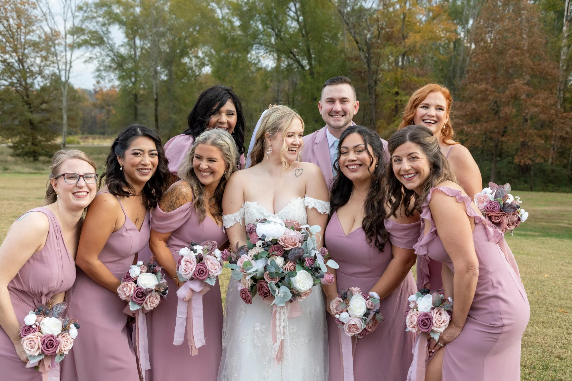 A group of women and one man gathered outdoors for a wedding photo, with colorful fall trees in the background. The women are dressed in matching mauve dresses, holding bouquets of pink, purple, and white flowers. The bride is in a white lace gown wi