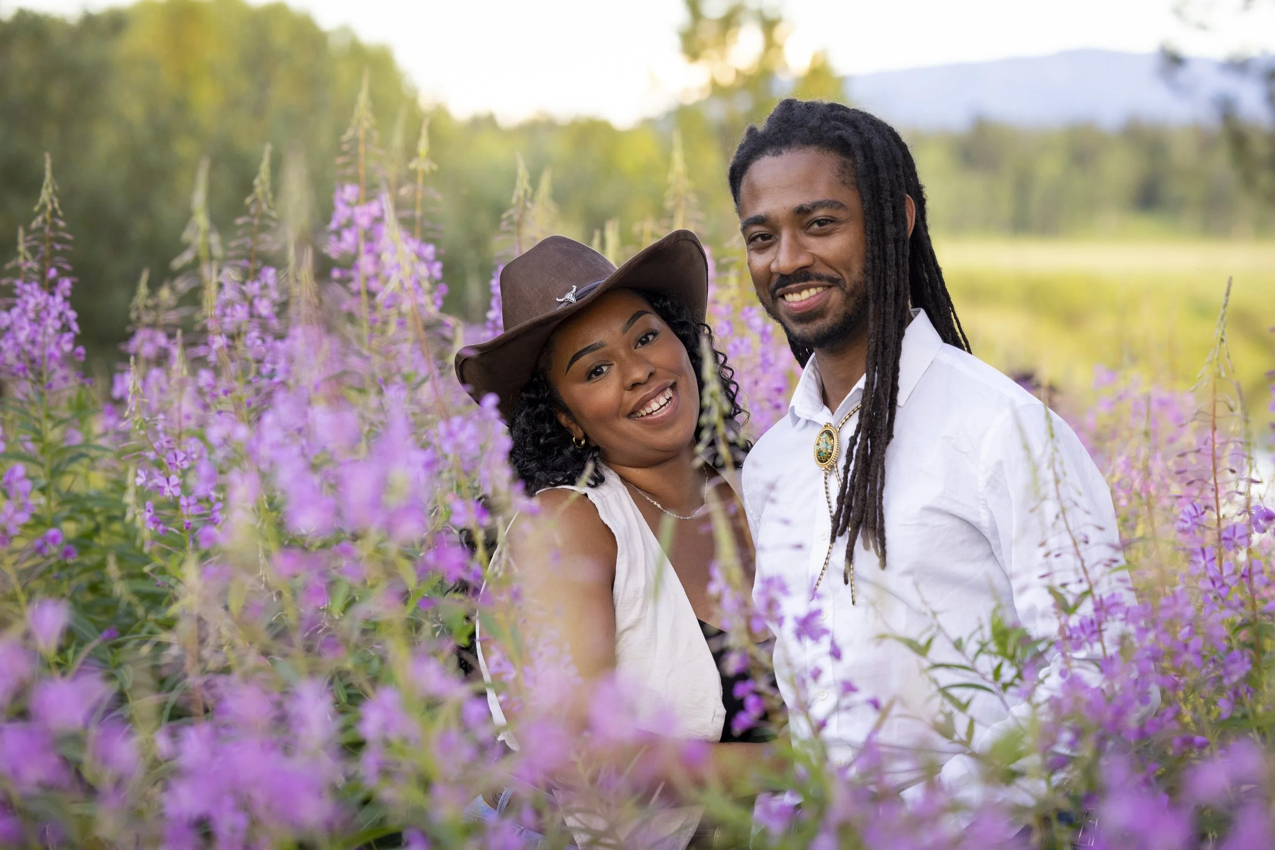 A smiling Black man with long dreadlocks and a white shirt, standing next to a smiling Black woman wearing a brown wide-brimmed hat with a flower detail, surrounded by purple wildflowers in a natural outdoor setting.