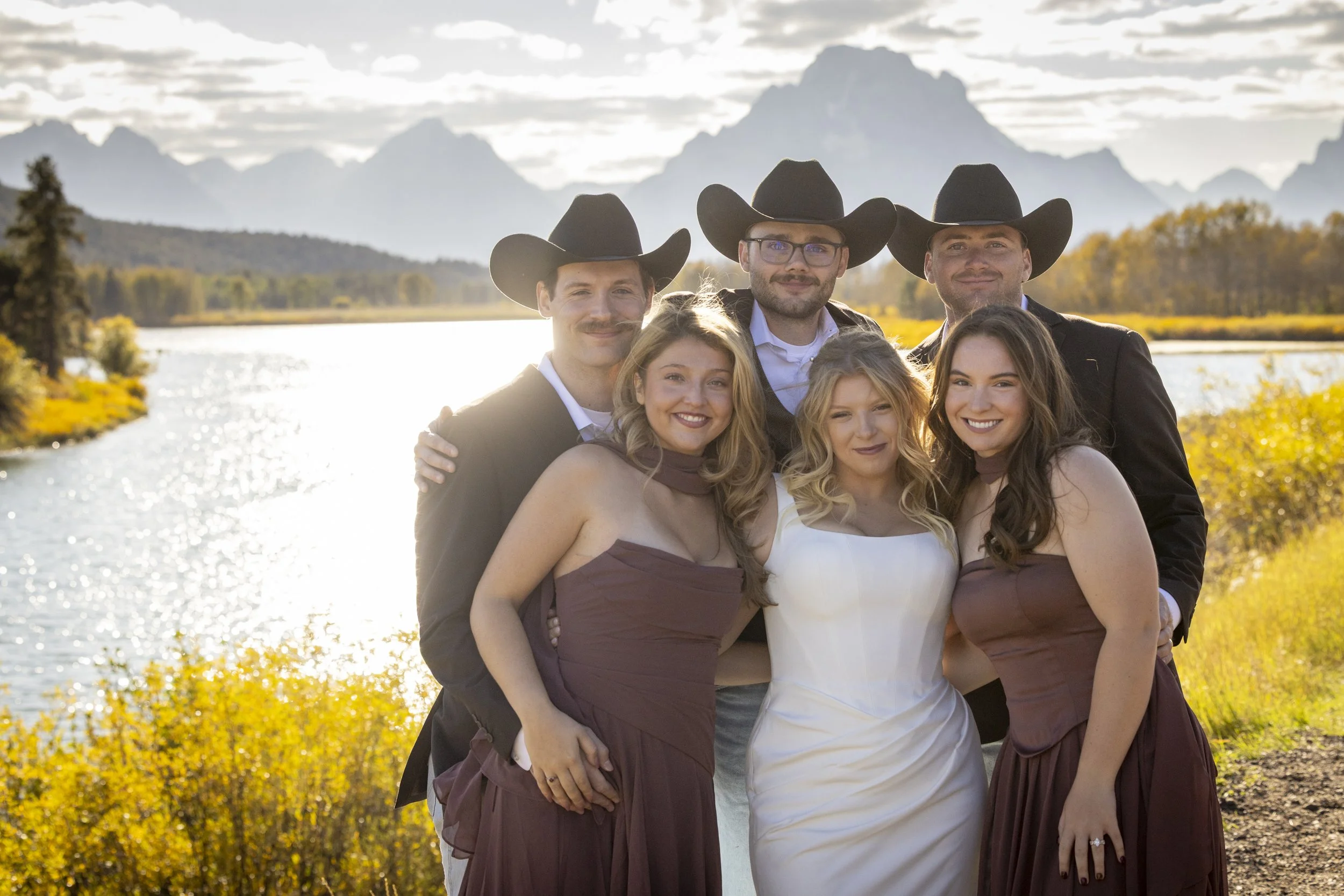 A group of six friends dressed in Western attire, wearing cowboy hats, standing outdoors near a body of water with mountains and trees in the background during sunset.